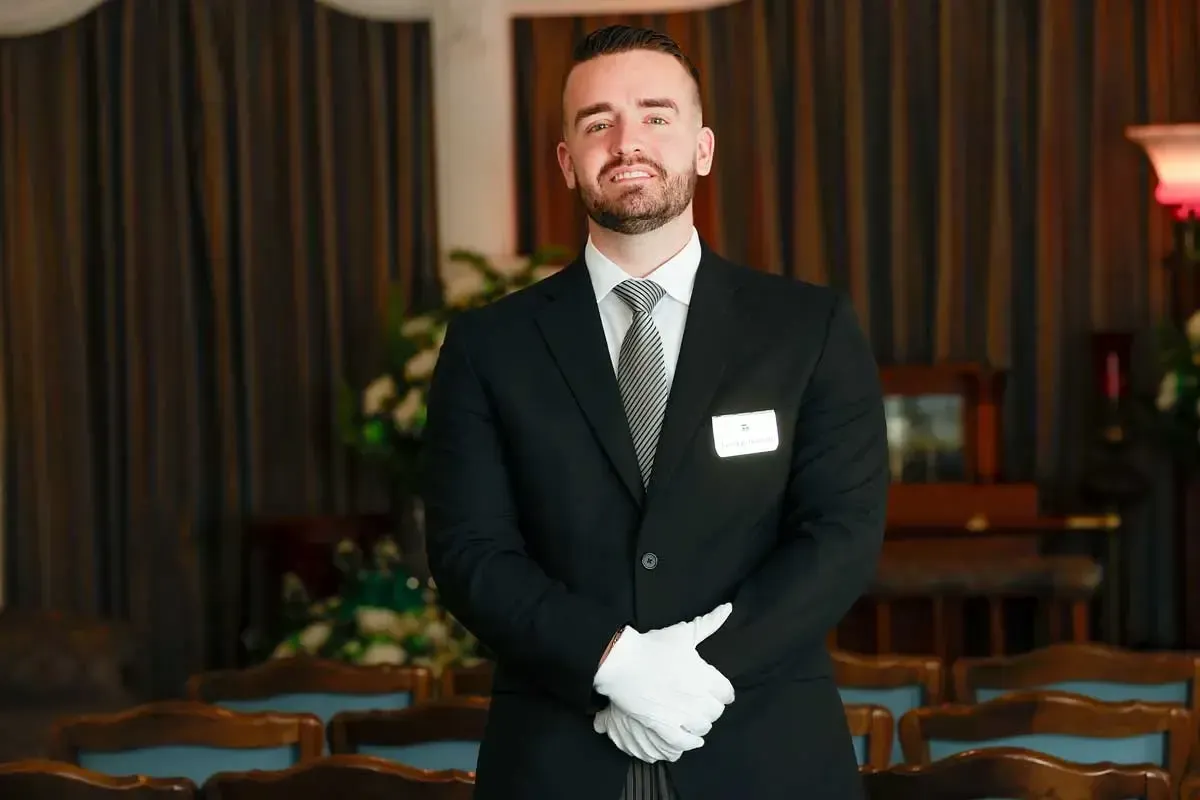Man in a black suit with white gloves standing in a formal room with curtains and chairs