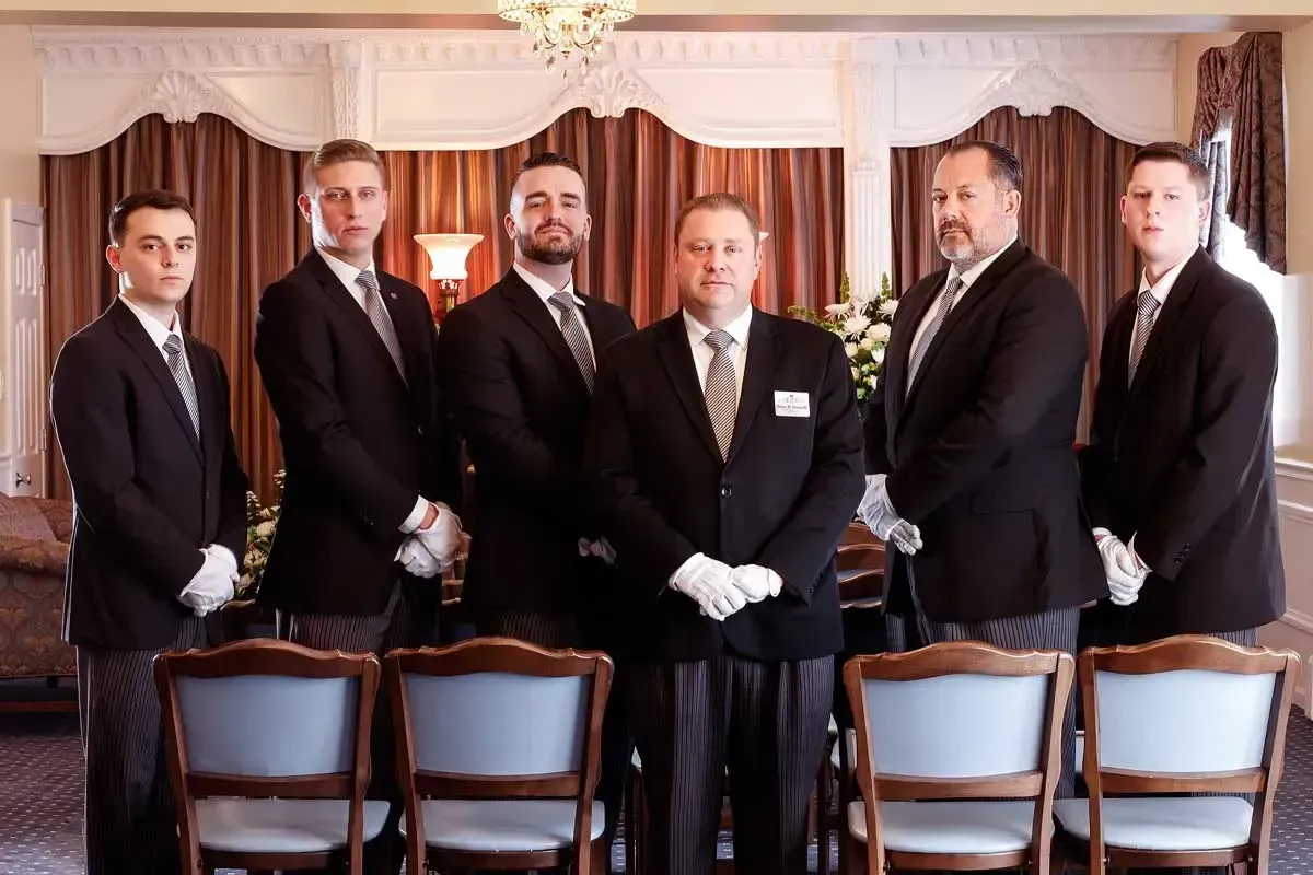 Six men in suits standing in a formal dining room, with blue-backed chairs in the foreground.