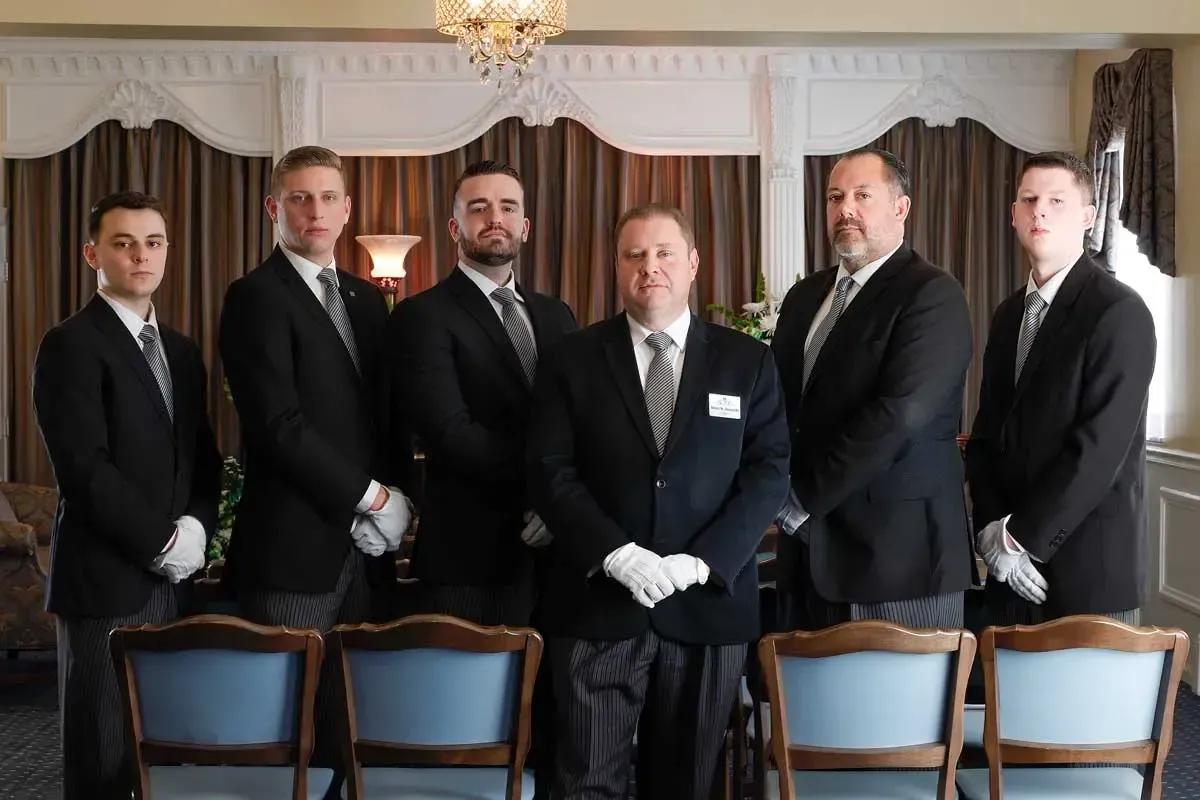 Six men in black suits and white gloves standing in a formal room with chairs in front of them