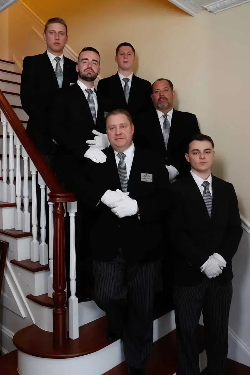 Six men in black tuxedos and white gloves pose on a staircase indoors.