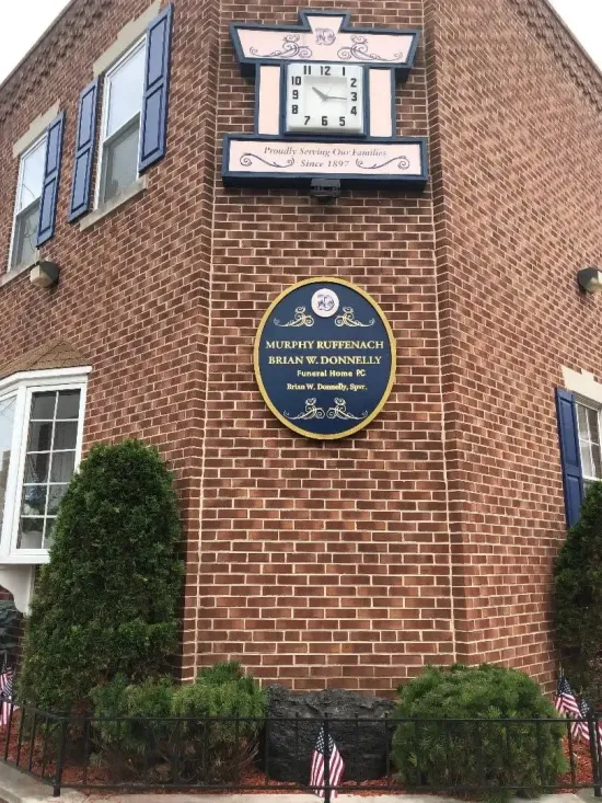 Brick building corner with historic plaques and a white-framed clock above a blue sign.