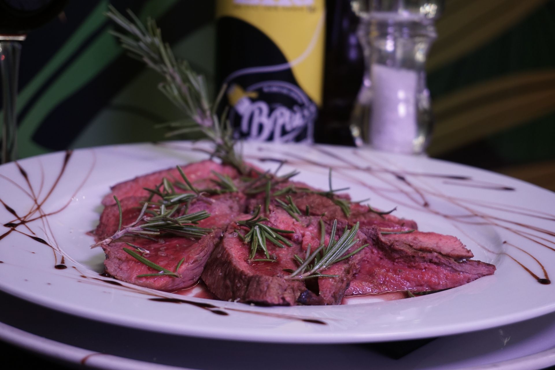 Un piatto bianco con sopra carne e rosmarino accanto a una bottiglia di birra.