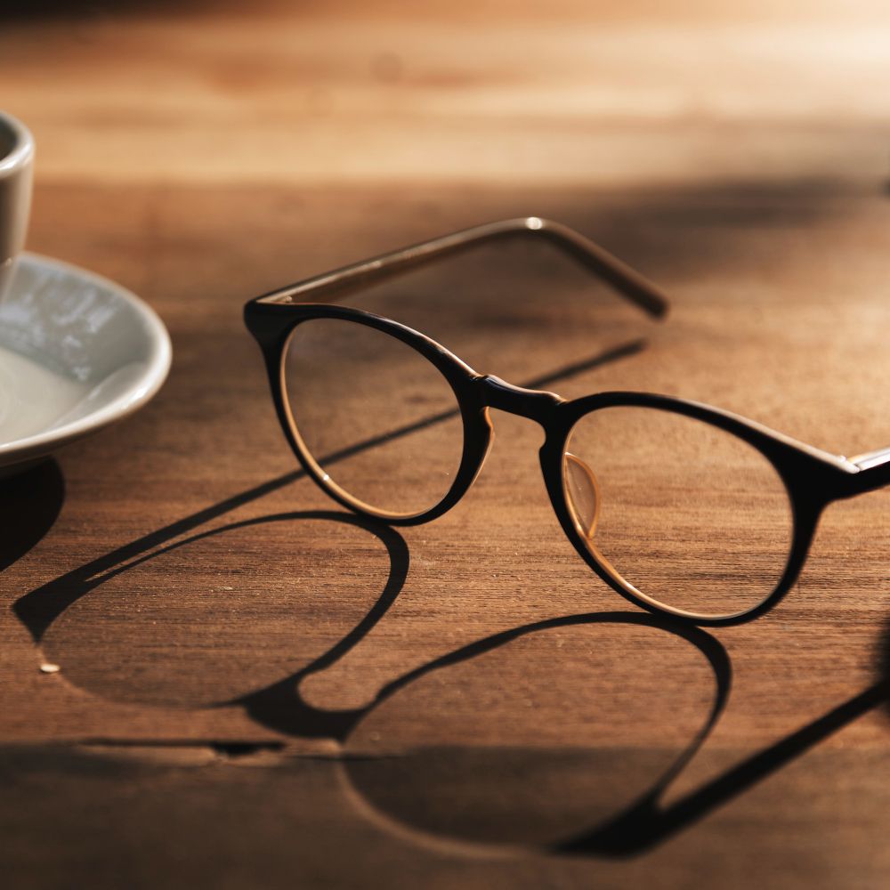Round black eyeglasses on a wooden table beside a coffee cup and saucer