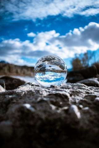 Glass sphere reflecting a blue sky and clouds on a rocky ground with blurred hills behind