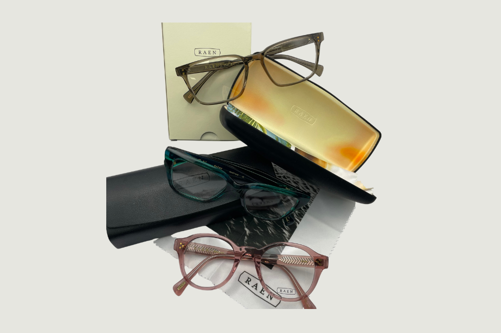 Three eyeglass frames displayed on a white surface beside a black-and-white eyeglasses box.