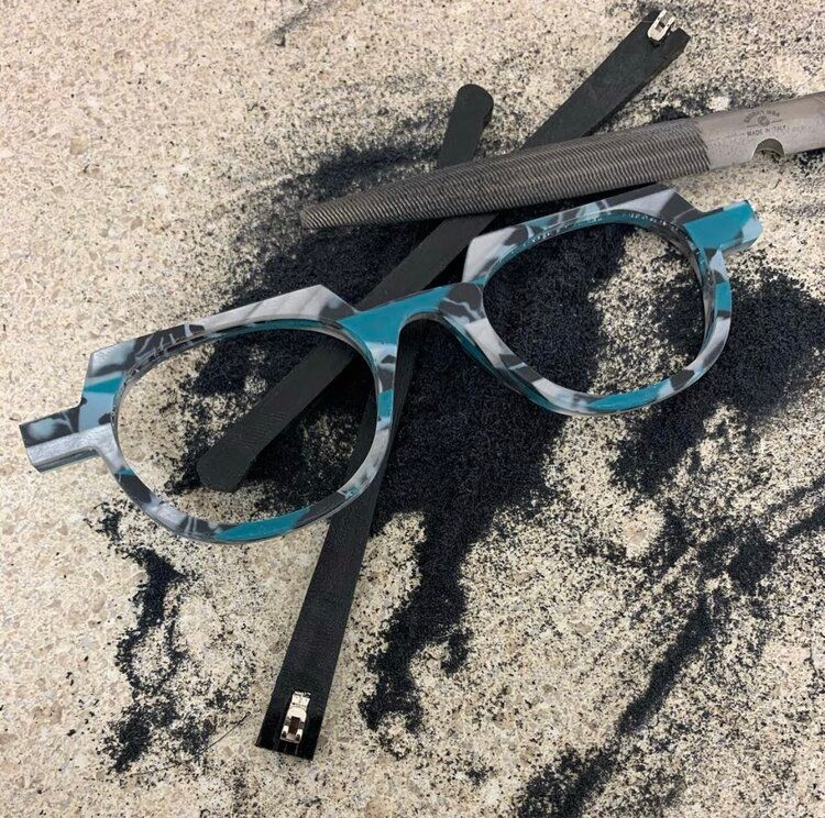 Blue-and-black eyeglasses lying on a sandy surface, with a black strap behind them.