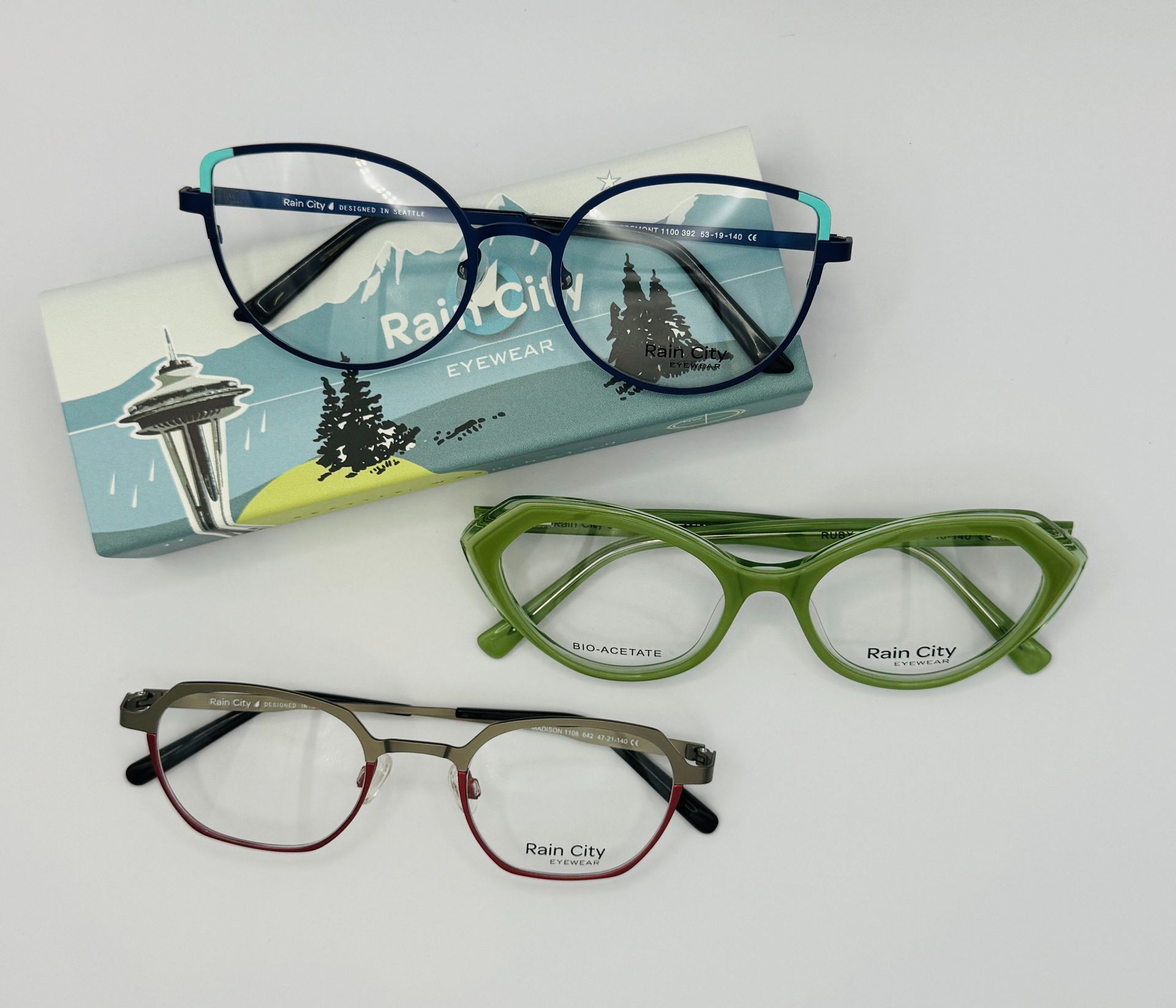 Three eyeglass frames on a Ray-Ban case: blue, green, and red-black.