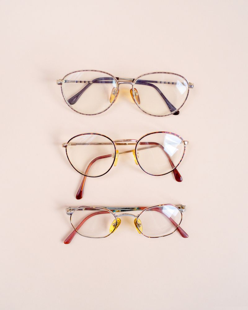 Three pairs of eyeglasses stacked vertically on a light background, in silver, gold, and red frames