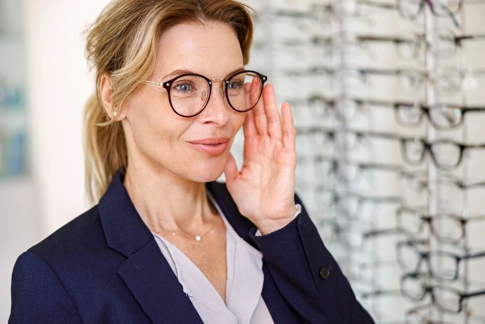Woman in glasses in a navy blazer, smiling and cupping her hand near her ear in an office setting