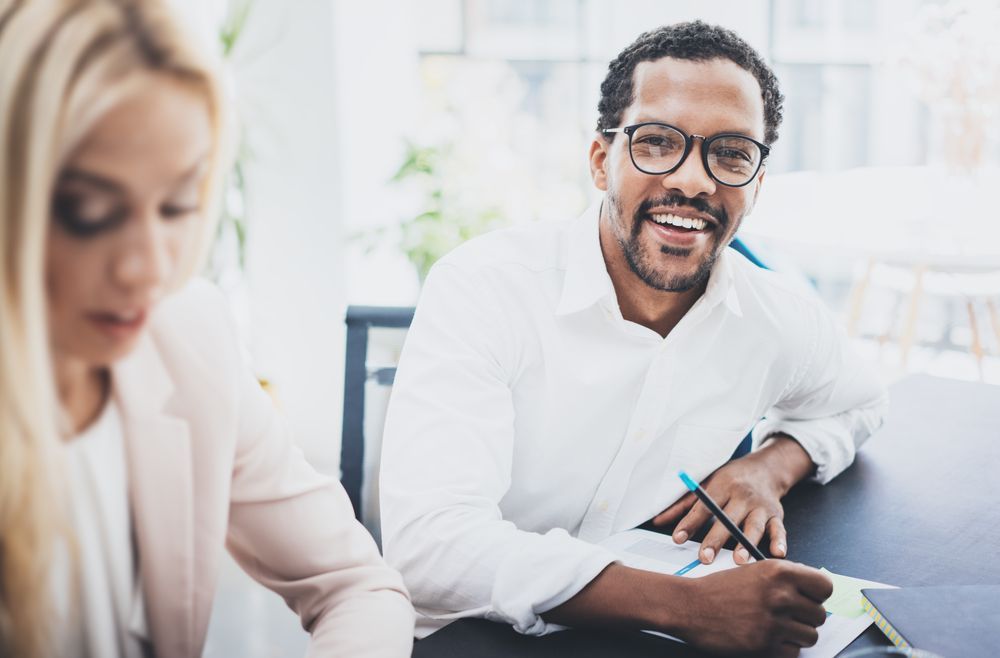 Two coworkers smiling at a desk, one writing on paper in a bright office