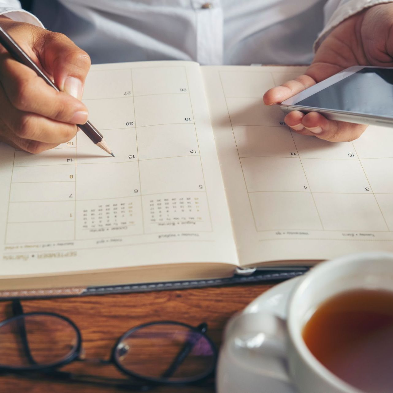 A person is writing in a planner while holding a cell phone