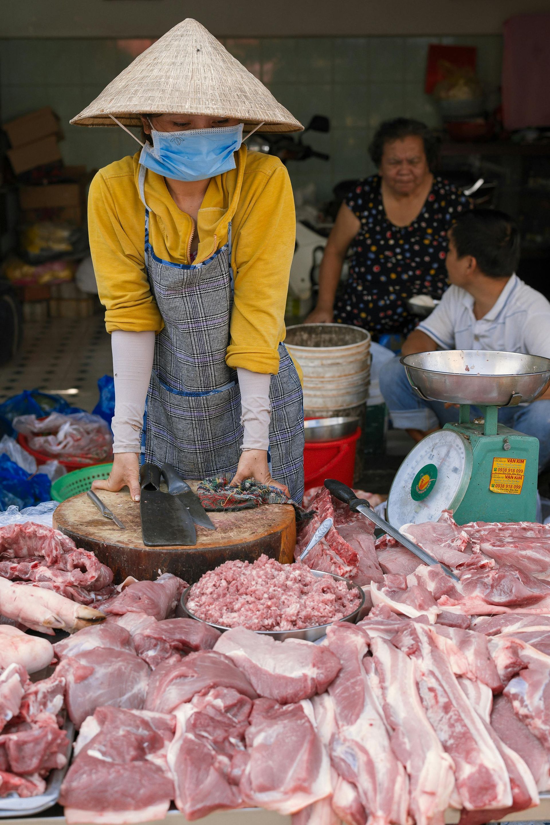 A person wearing a conical hat and face mask works at a butcher stand filled with cuts of raw meat at an open-air market.