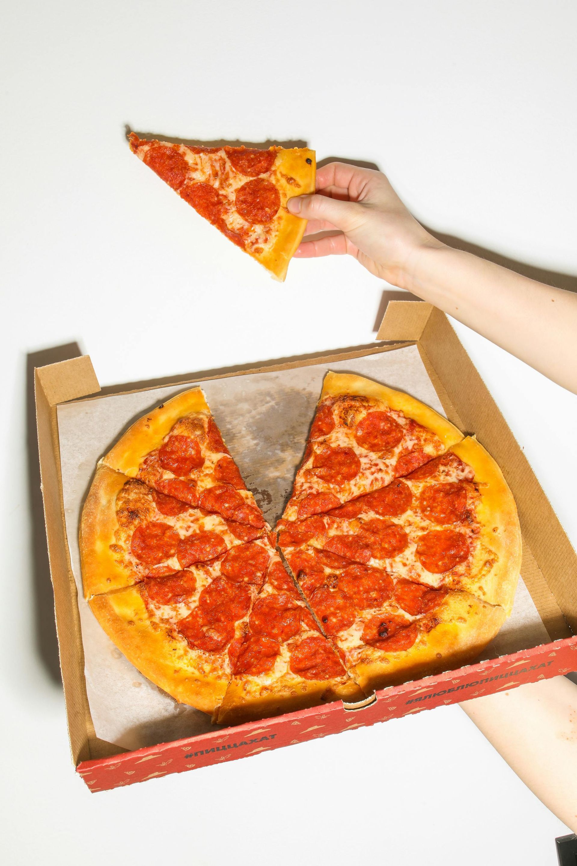 A hand lifts a slice of pepperoni pizza from a cardboard box containing the rest of the pie.