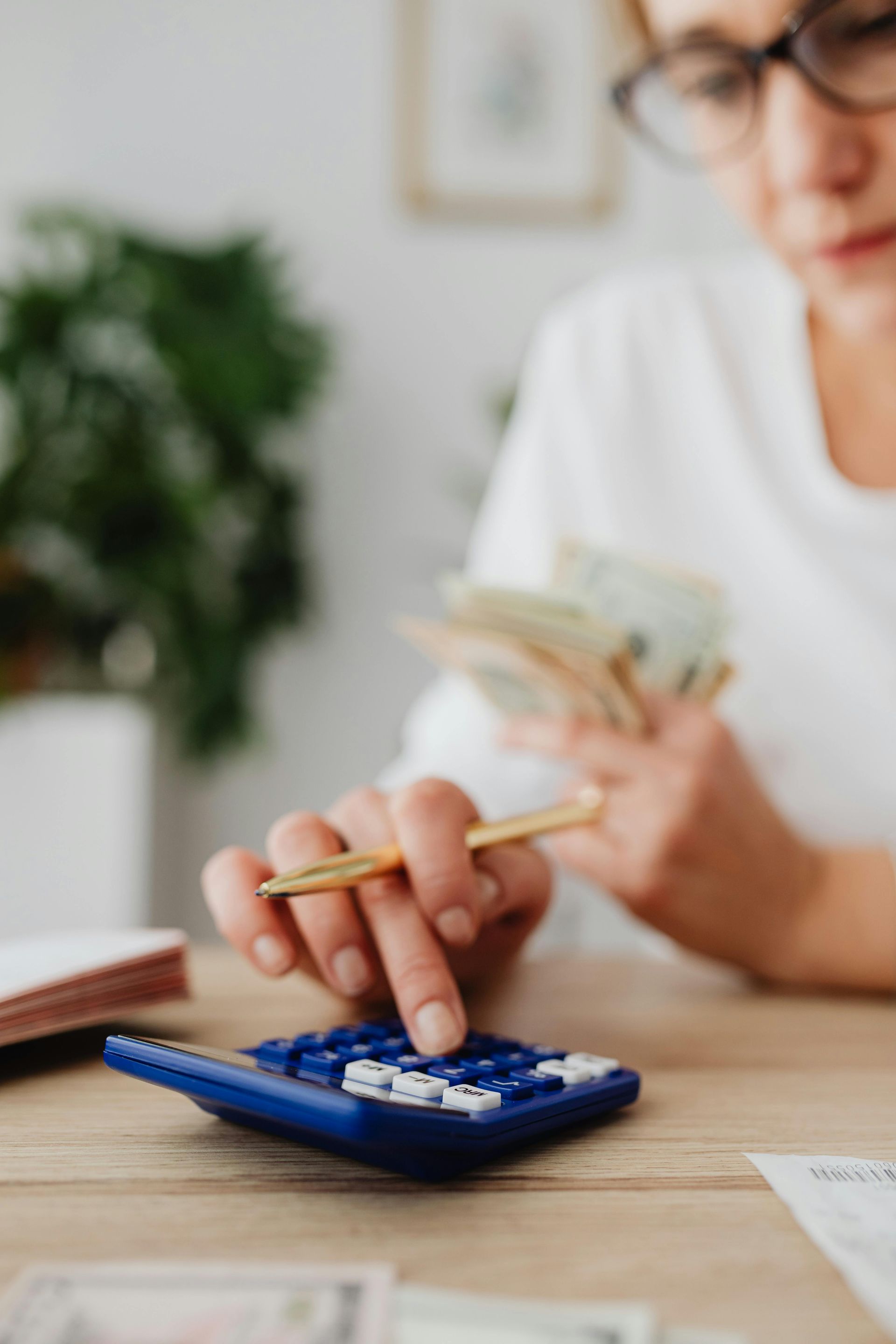 A person with glasses sits at a desk, holding cash while calculating numbers on a blue calculator.