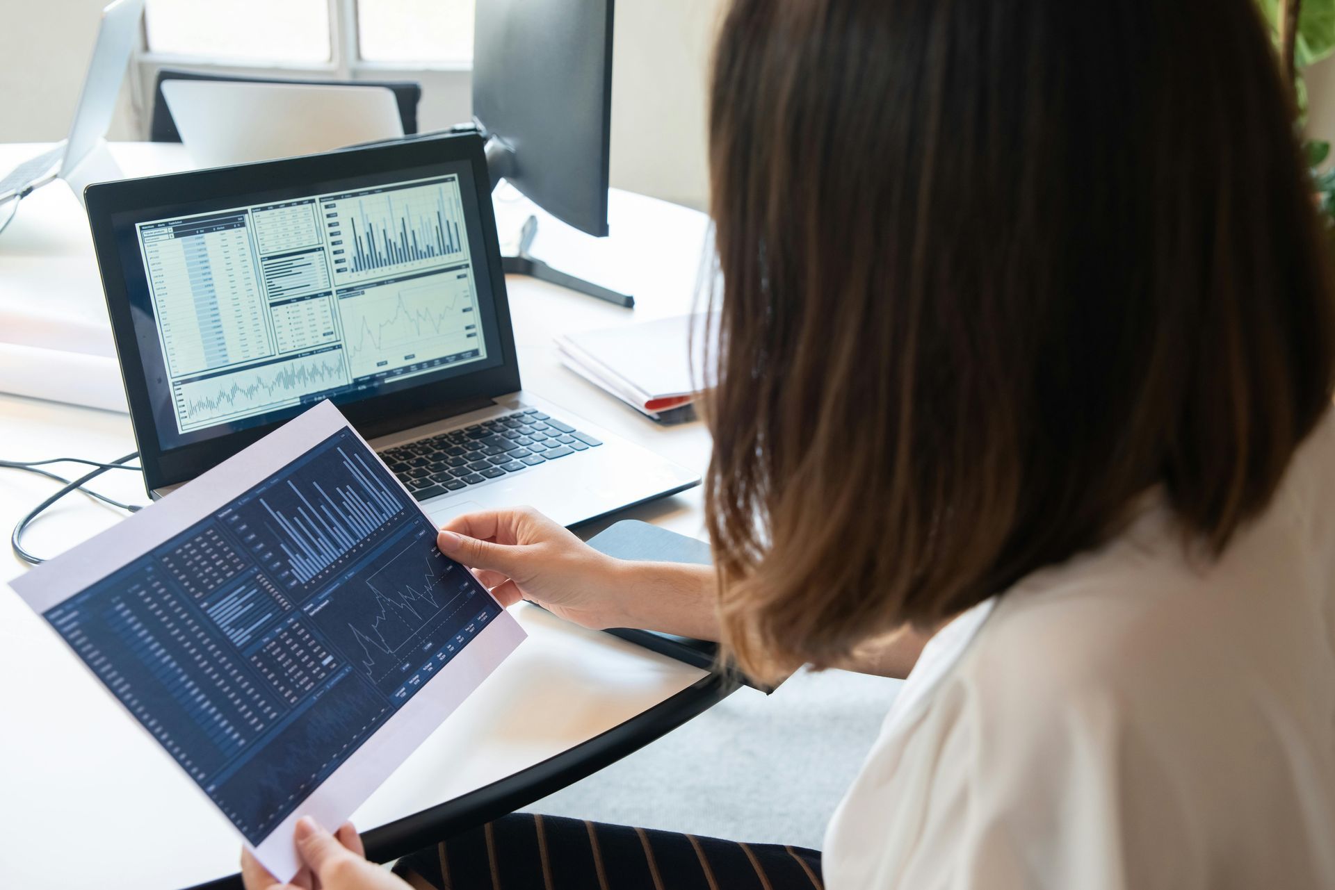 A person in an office holds a printed chart, comparing it to data displayed on a laptop screen.
