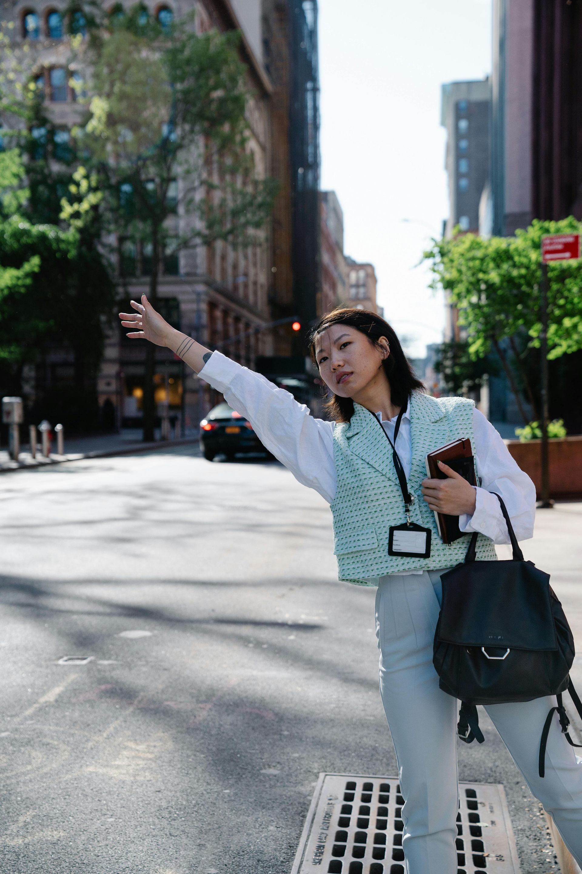 A person in a light blue patterned vest hails a taxi on a city street while carrying a black backpack.