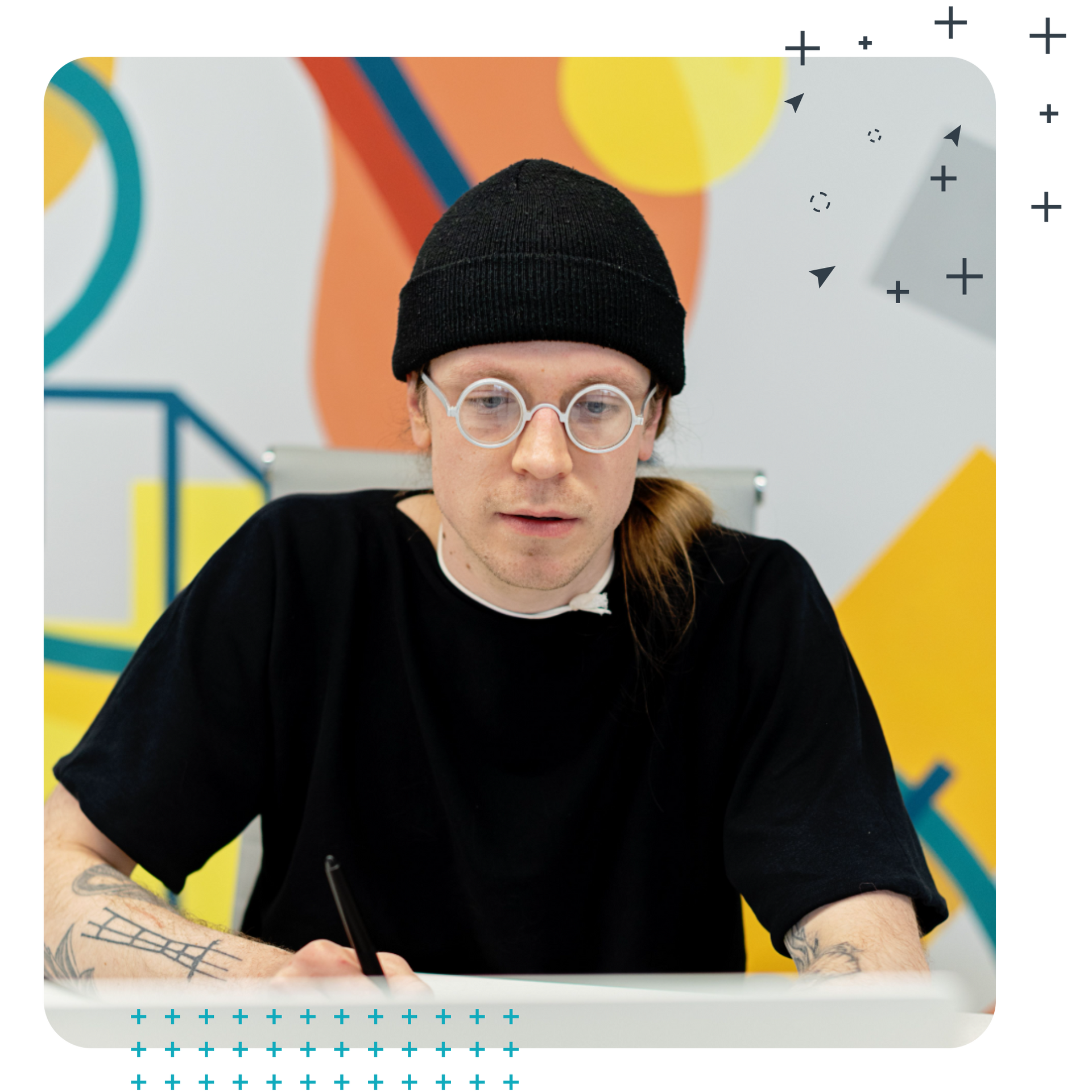 A person in a black beanie and glasses writes at a desk against a colorful mural background in a gallery.