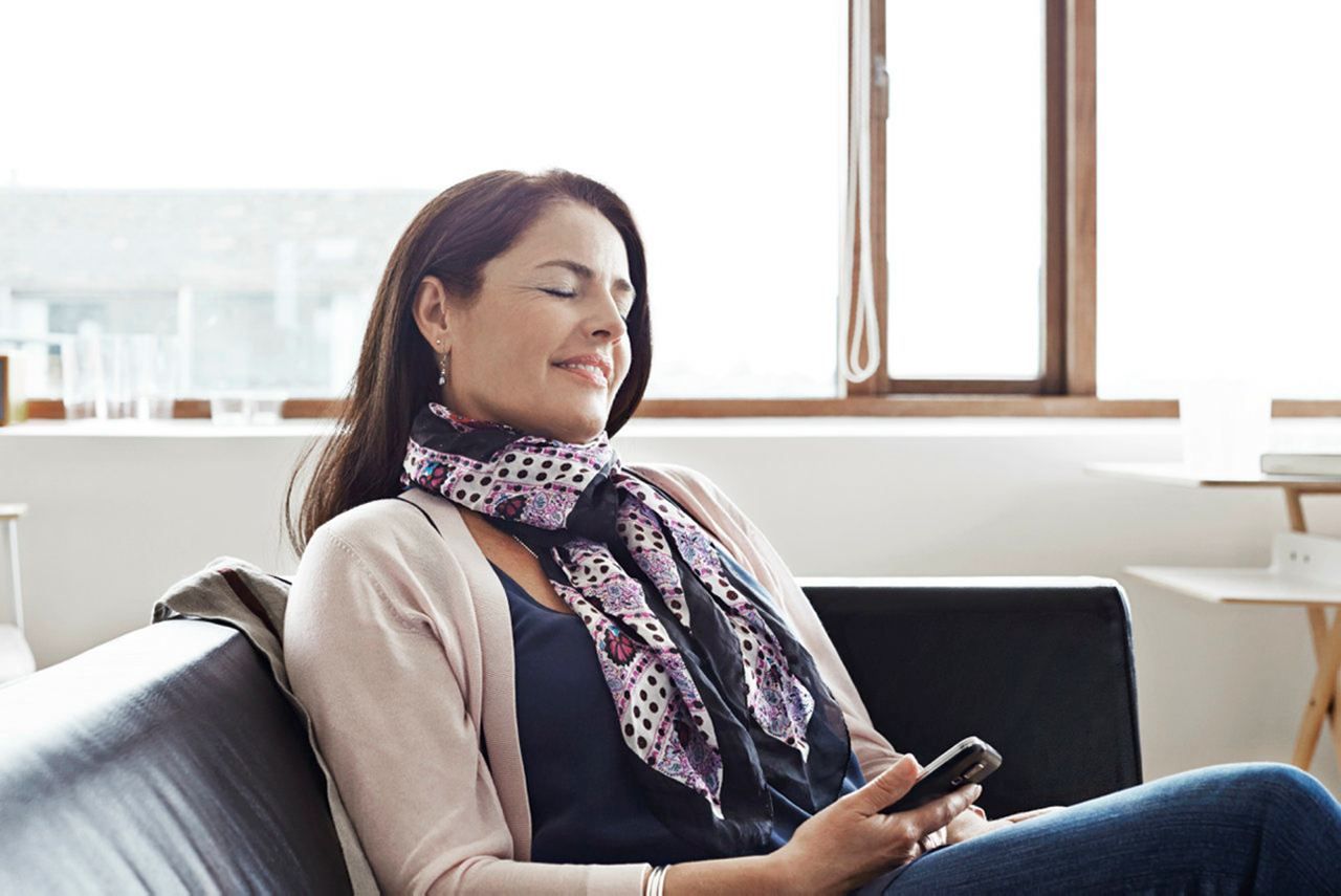 Woman relaxing on a couch, smiling with eyes closed and holding a phone by a bright window — Local Hearing Aid Specialists in Wallsend, NSW