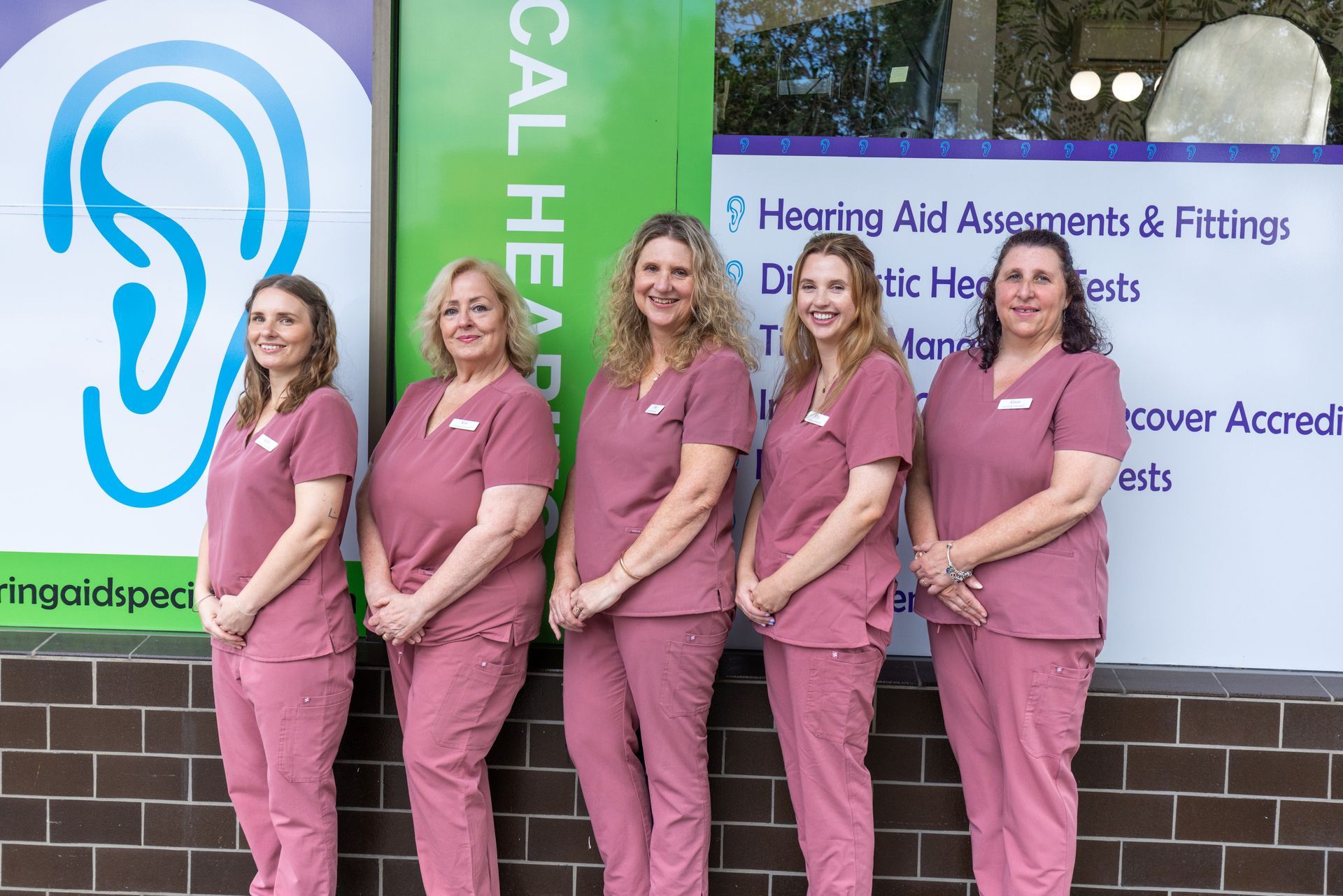 Five hearing clinic staff in pink scrubs stand smiling outside a clinic sign — Local Hearing Aid Specialists in Wallsend, NSW