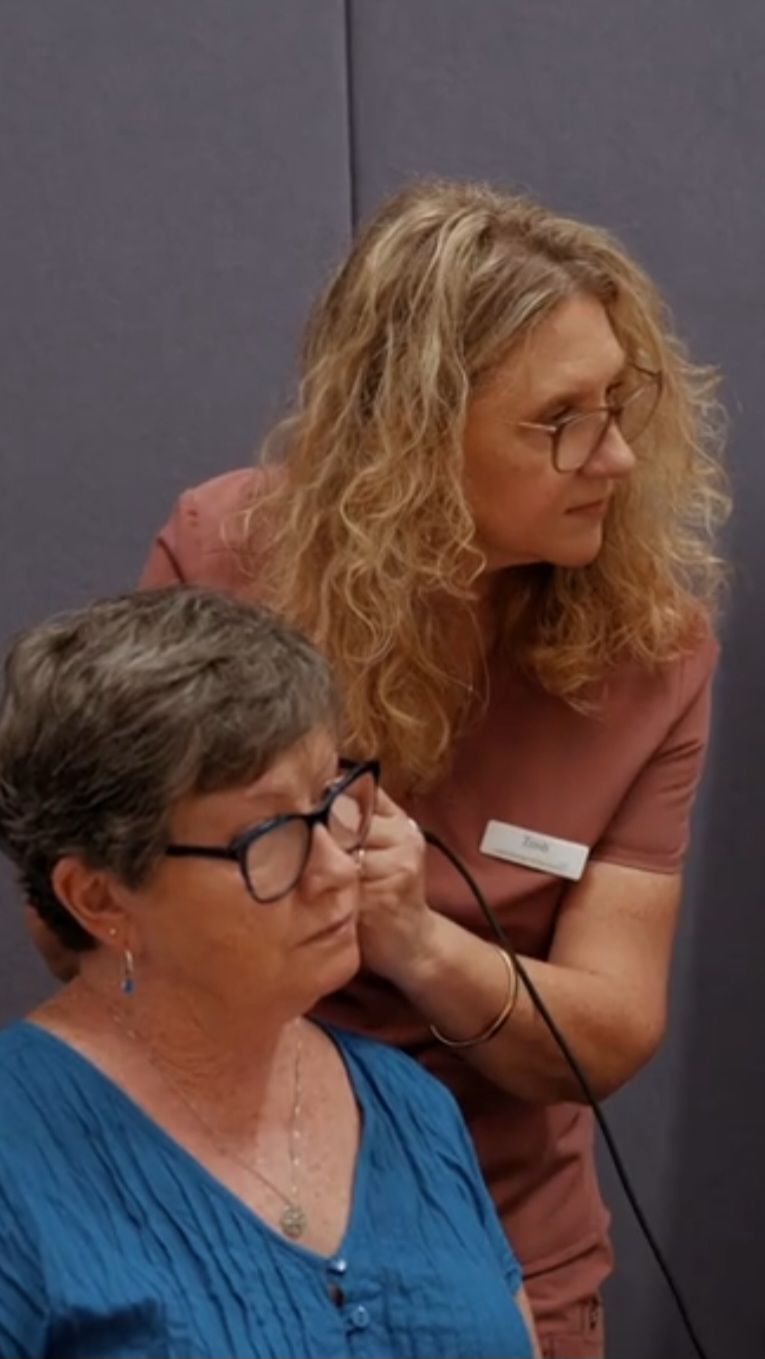 Two women seated indoors, one leaning in and speaking while the other looks ahead in blue and glasses — Local Hearing Aid Specialists in Wallsend, NSW