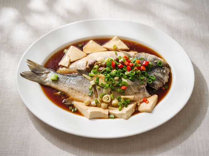 Whole steamed fish on a white plate with soy sauce, tofu, and chopped green onions with red chilies