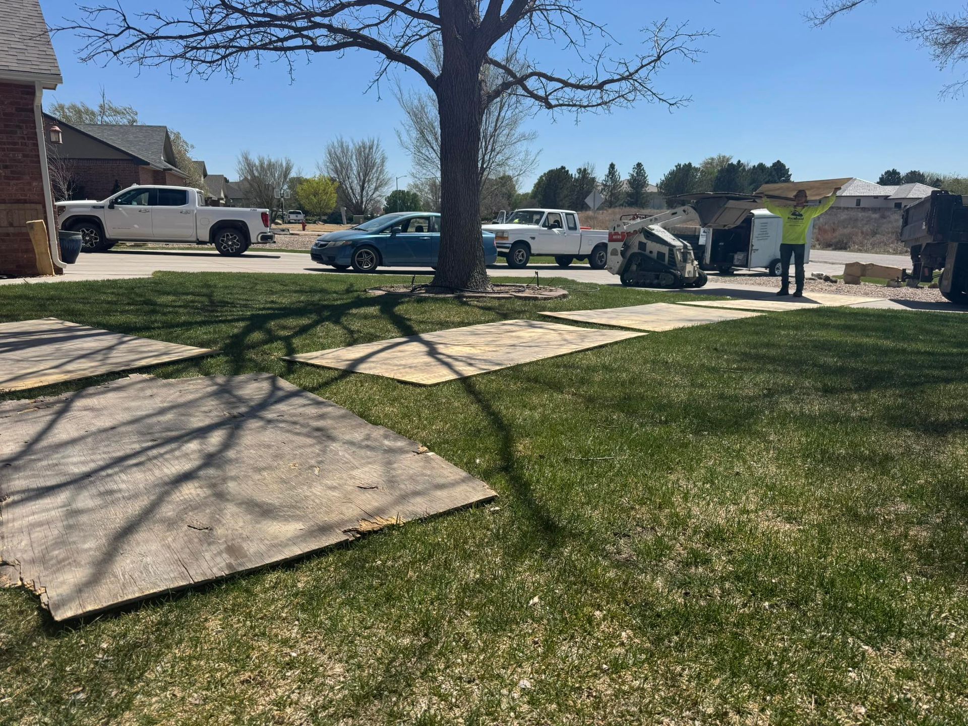 Suburban yard with two workers near a parked car, a white van, and a large tree casting shadows on the lawn.