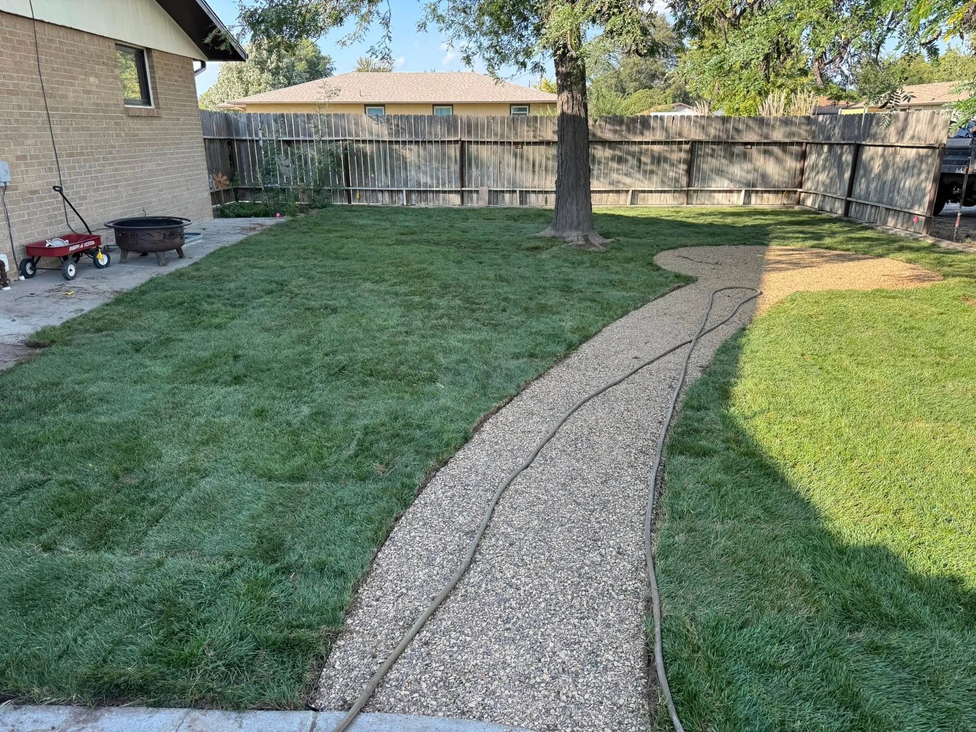 Backyard with a curved gravel path, green lawn, tree, wooden fence, and patio beside a house