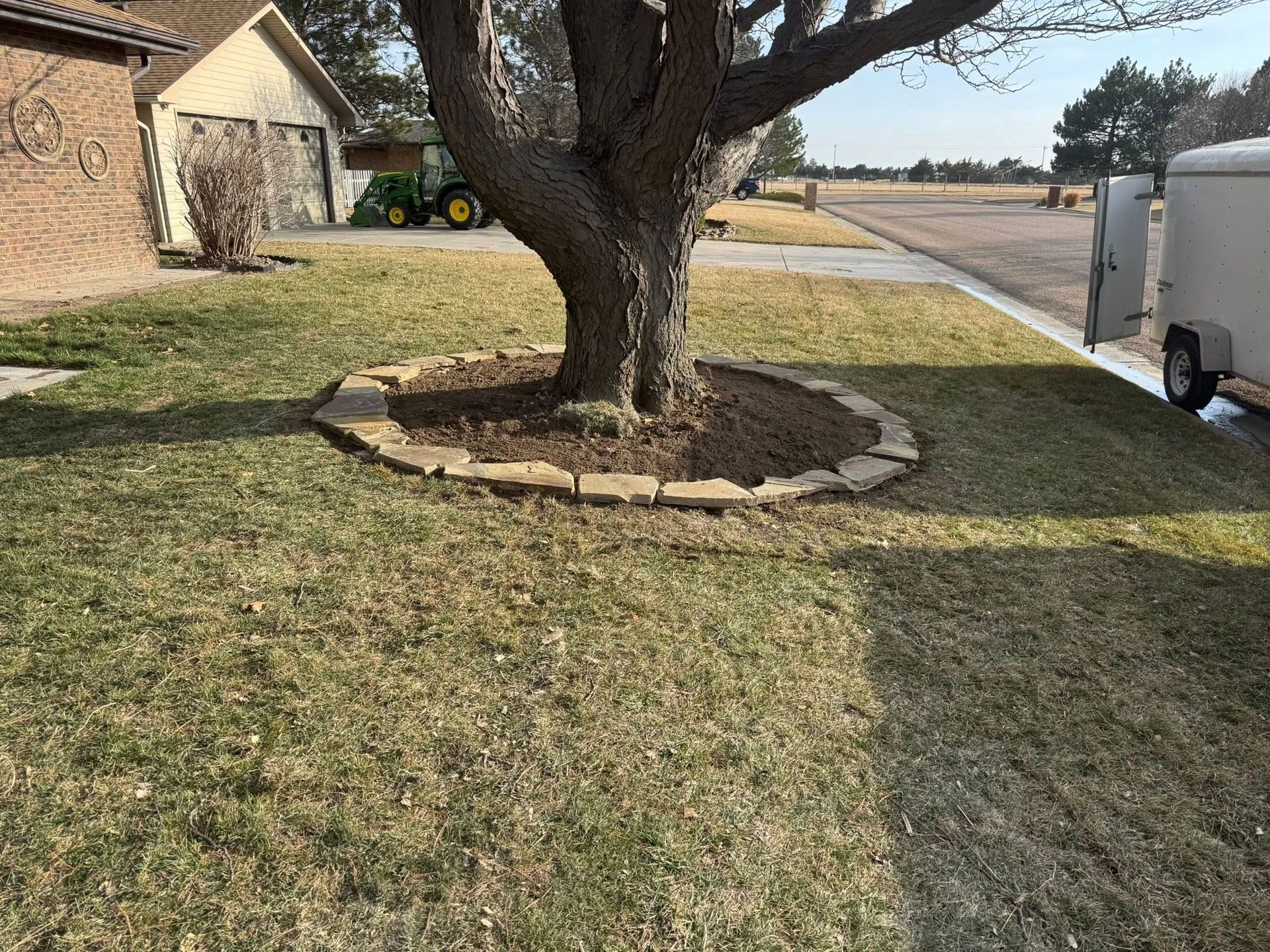 Front yard with a large tree surrounded by a stone border, next to a driveway and road in a suburban neighborhood