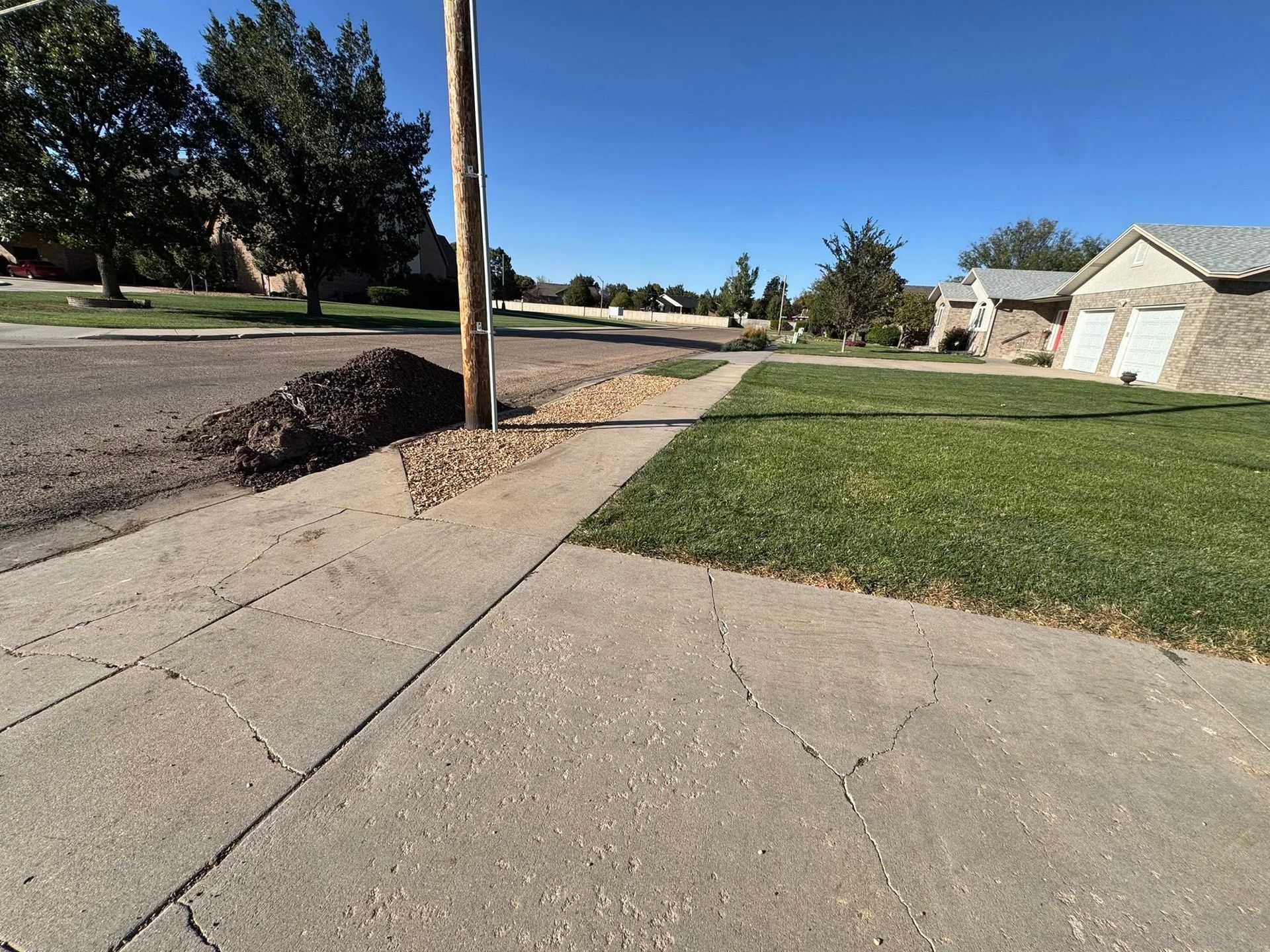 Suburban sidewalk beside a grassy lawn, street, and utility pole on a sunny day