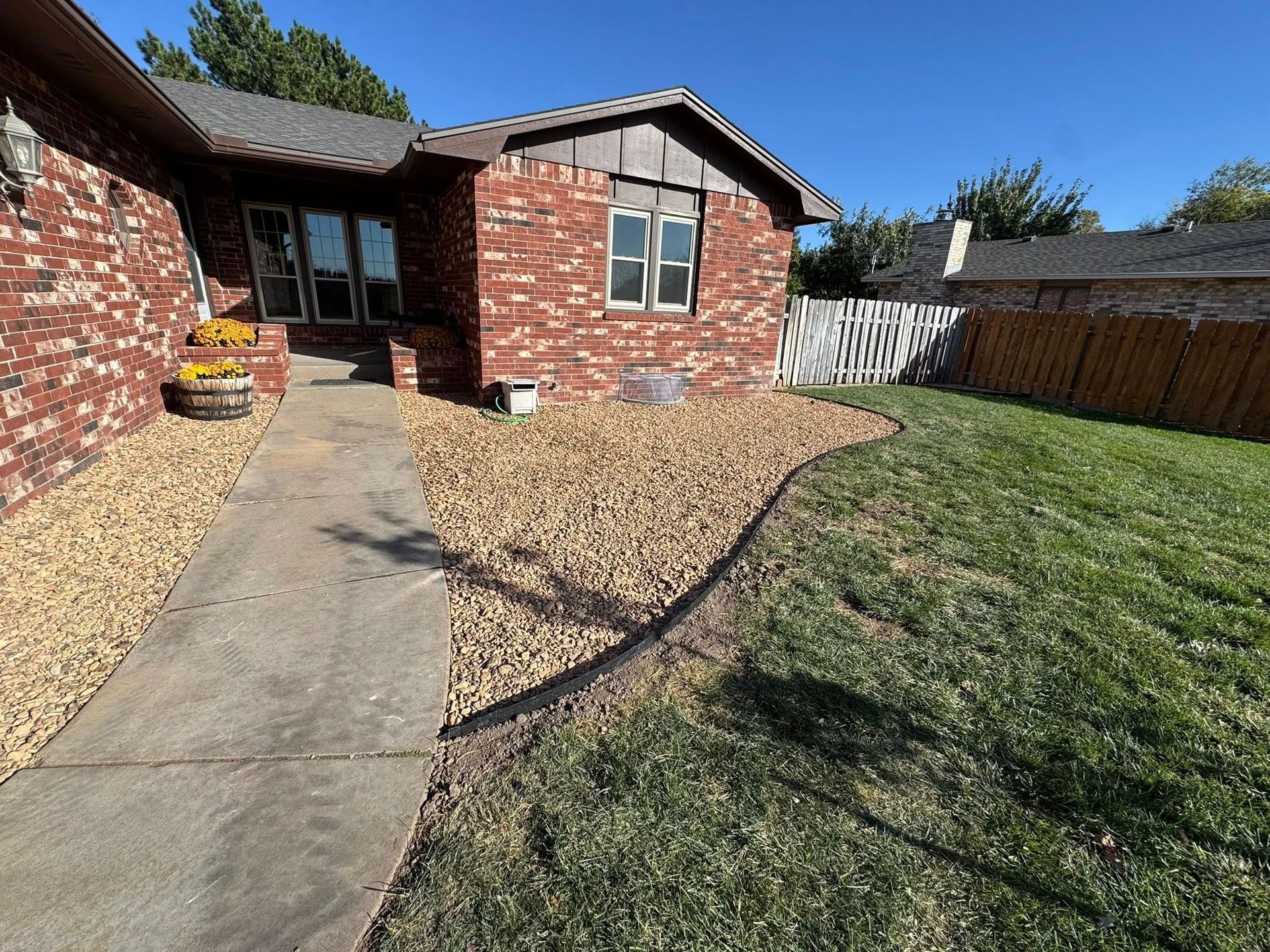 Backyard with brick house, concrete path, gravel area, and green lawn under clear blue sky