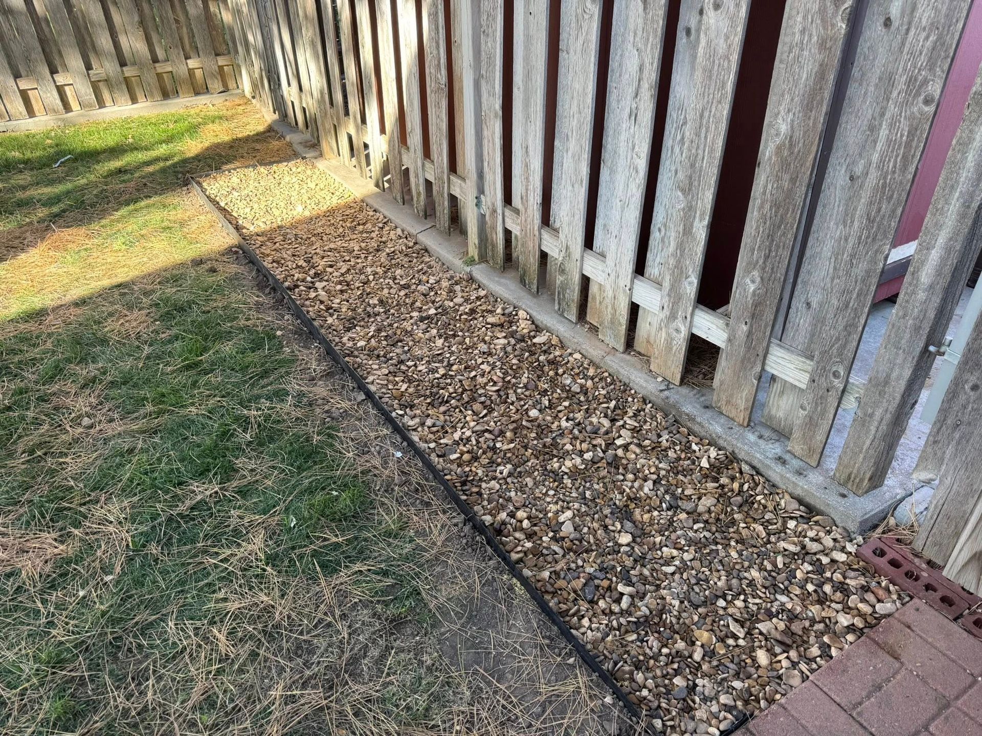 Gravel strip along a wooden fence beside a grassy yard, with sunlight and tree shadows.