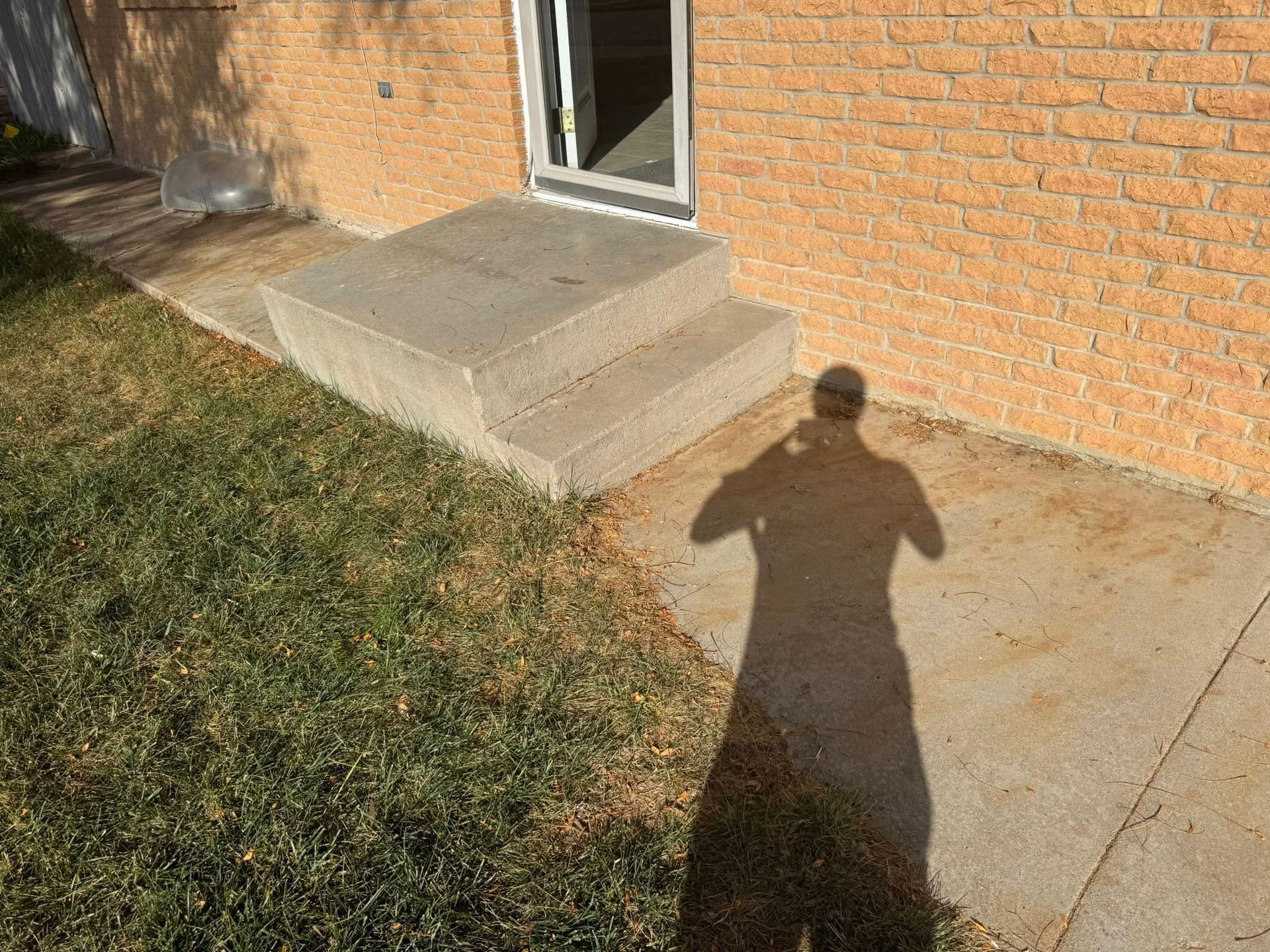 Concrete steps by a brick wall leading to a door, with a person’s shadow on the pavement and grass nearby