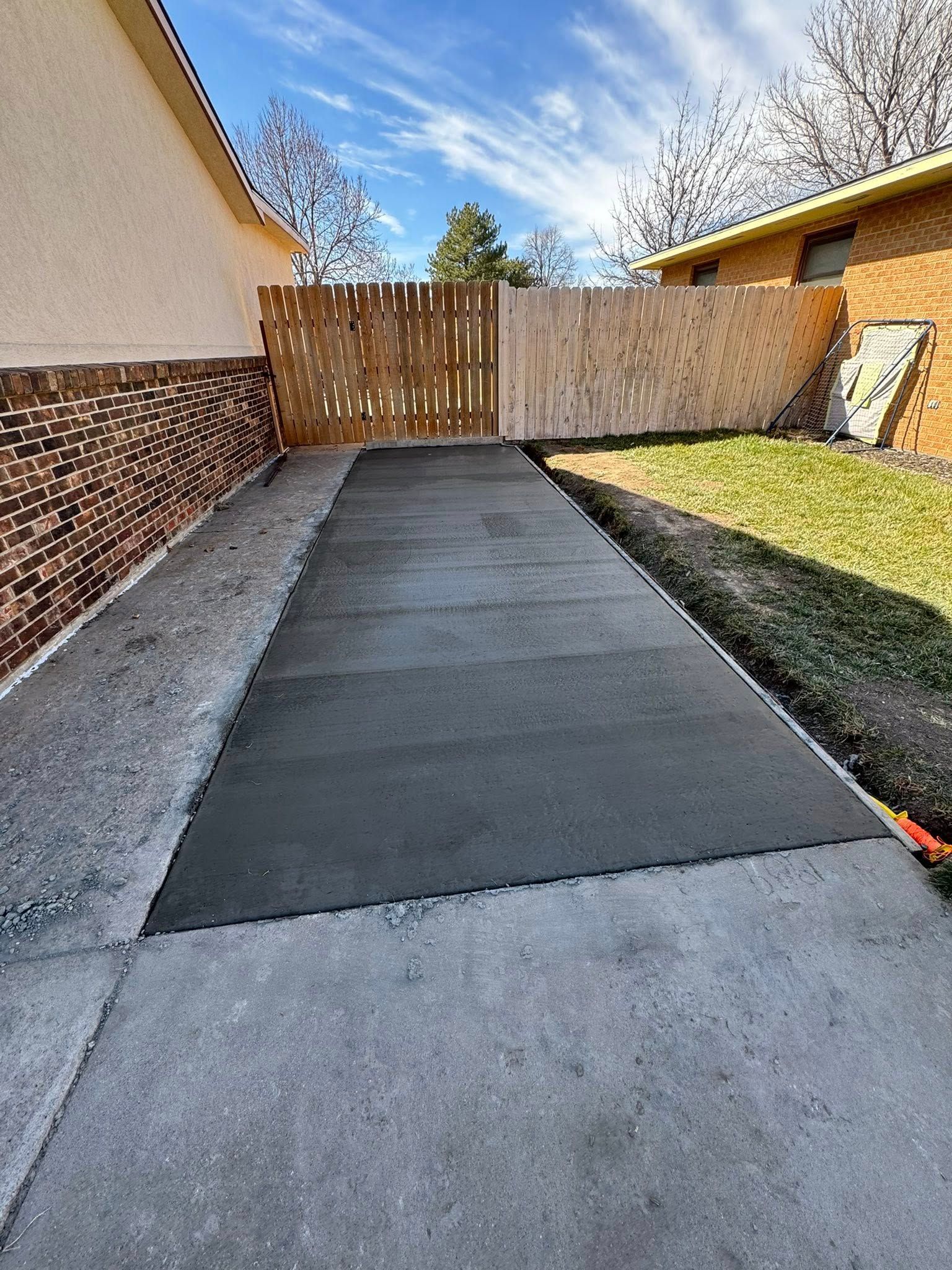 Concrete driveway with fresh dark patch leading to a wooden gate beside a house and grassy yard