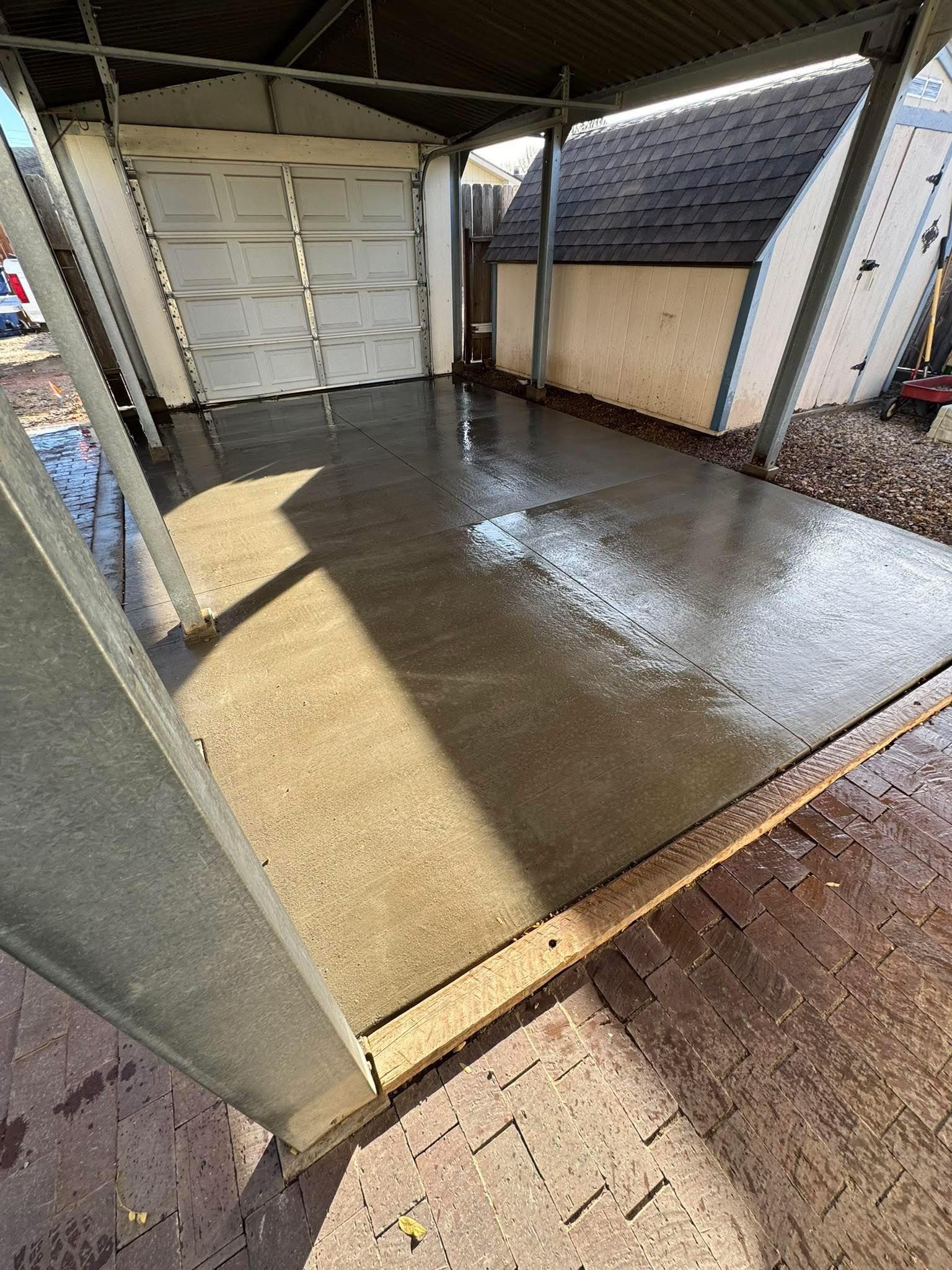 Wet concrete garage floor under a covered carport, with sunlight and a white garage door.