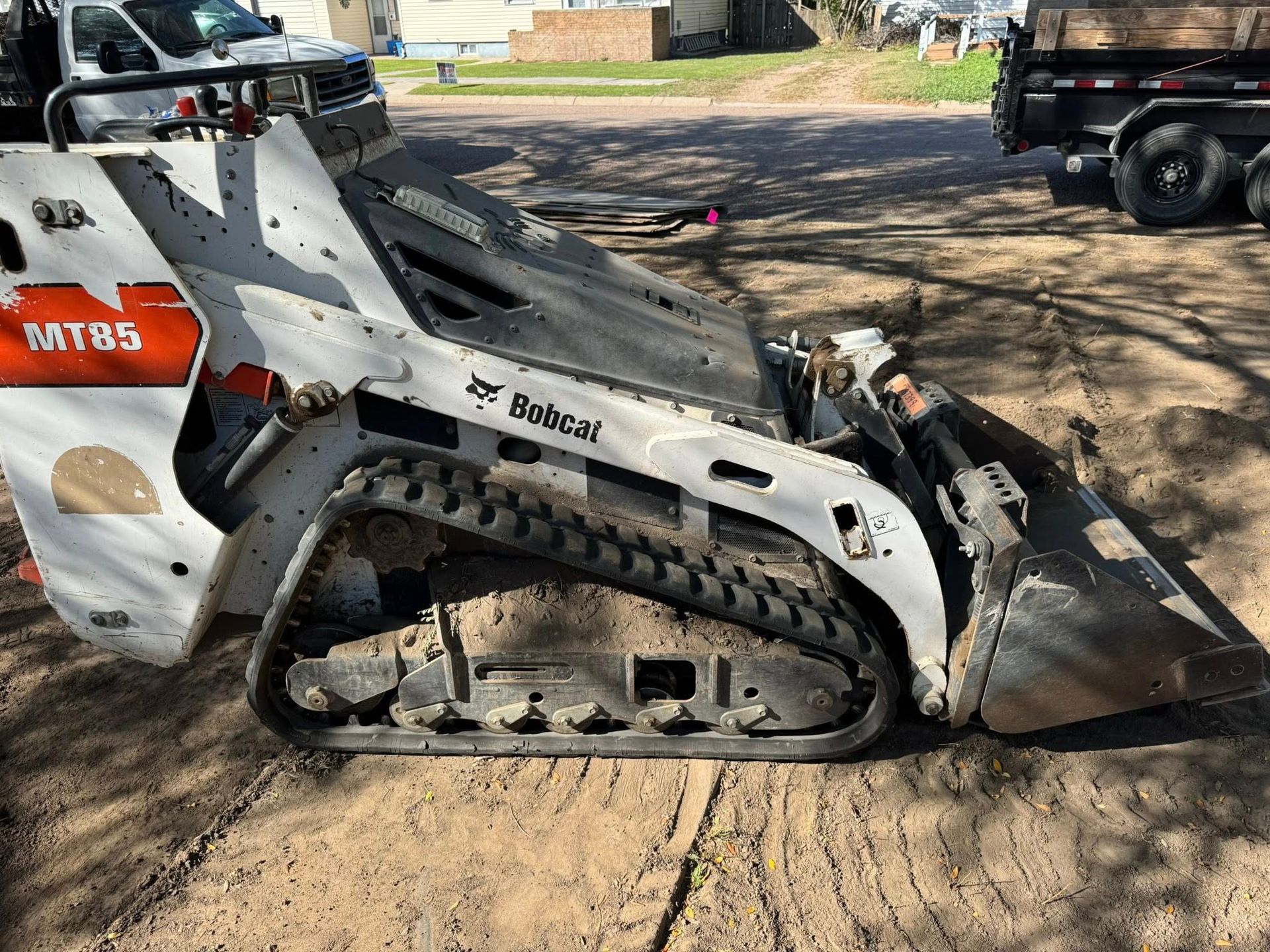 Compact skid steer loader with bucket parked on a dirt driveway in a wooded area.