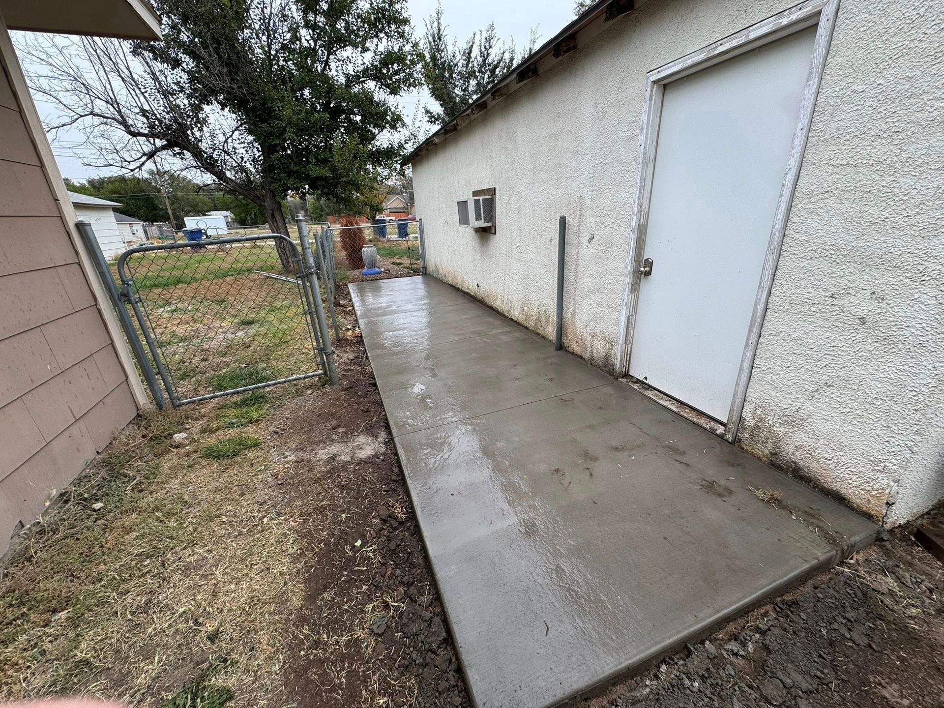 Narrow wet concrete walkway beside a white stucco building and chain-link fence after rain