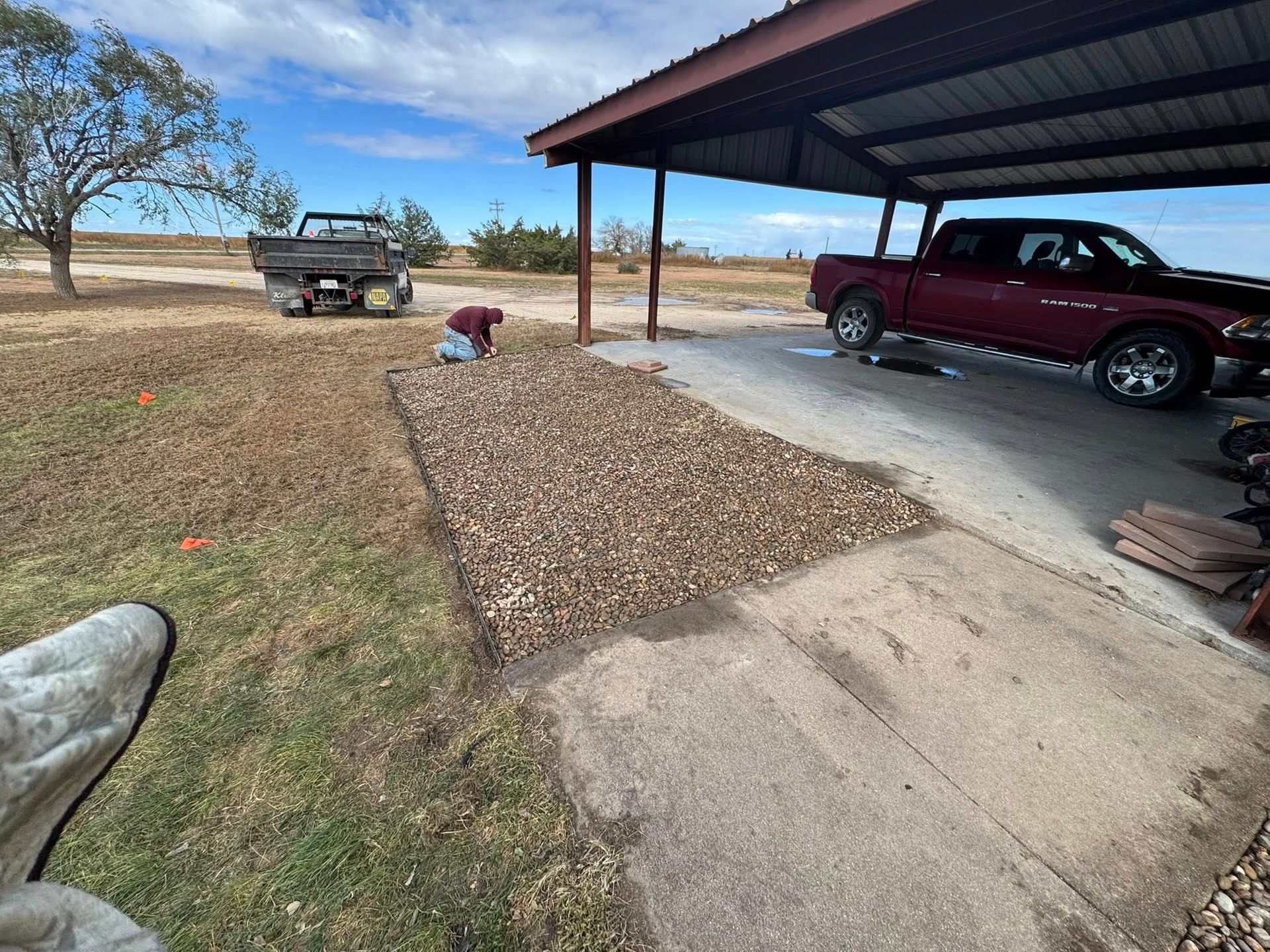Driveway with a red pickup under a carport, a gravel path, and a trailer in the yard under a blue sky.