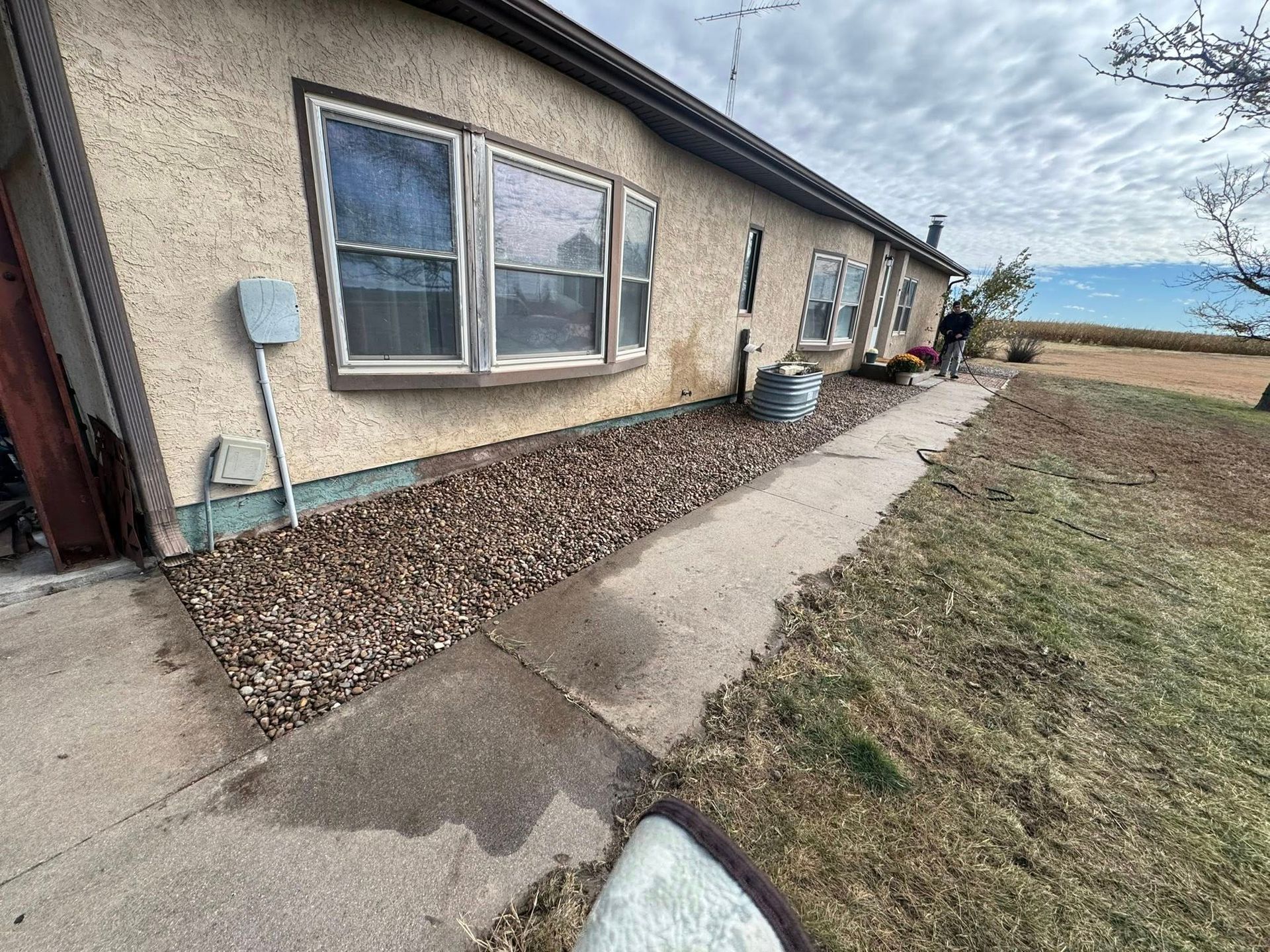 Narrow concrete walkway beside a beige house with gravel strip and open grassy yard under cloudy sky