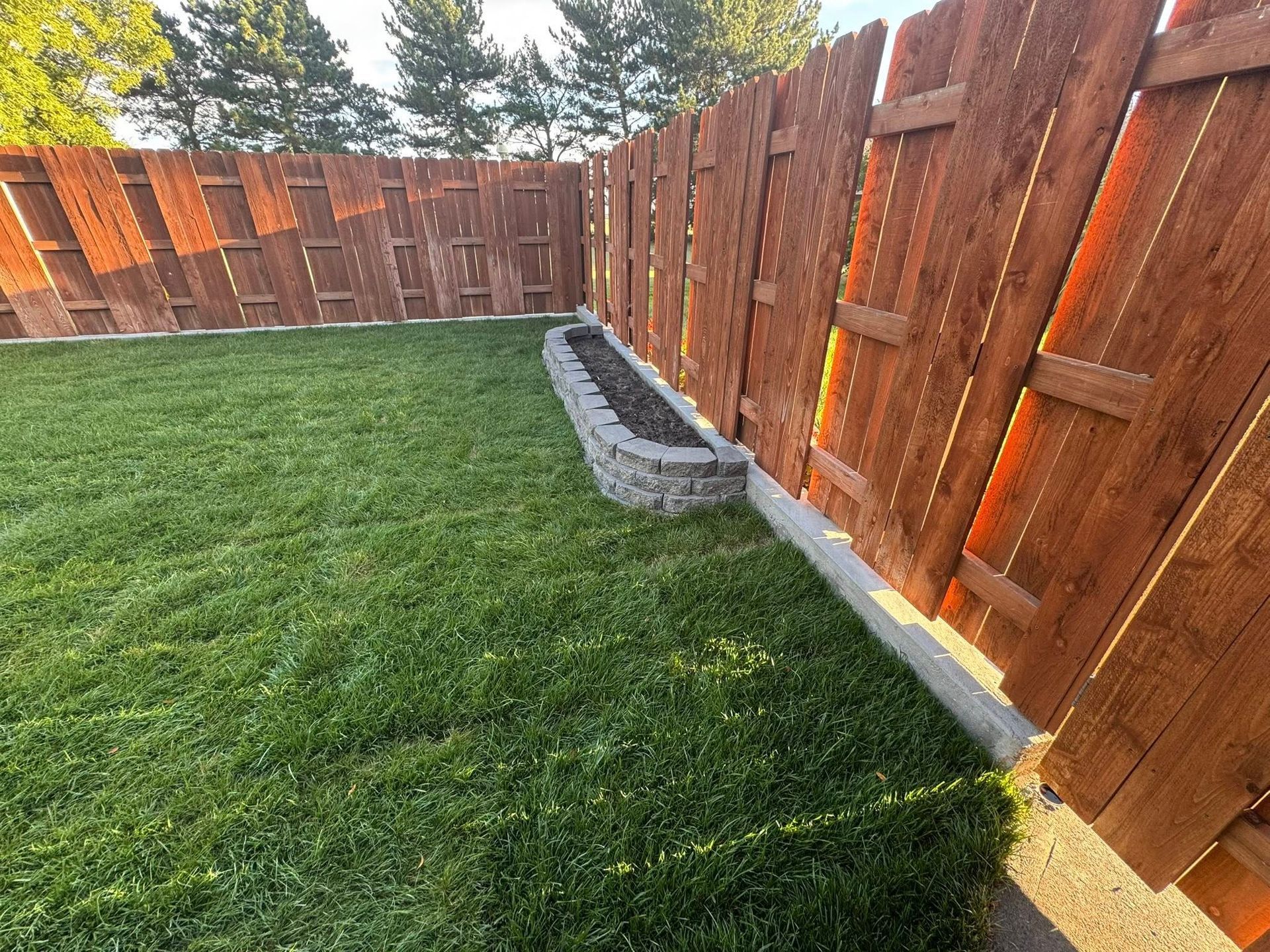 Backyard lawn beside a wooden fence with a small stone planter and trees in the background