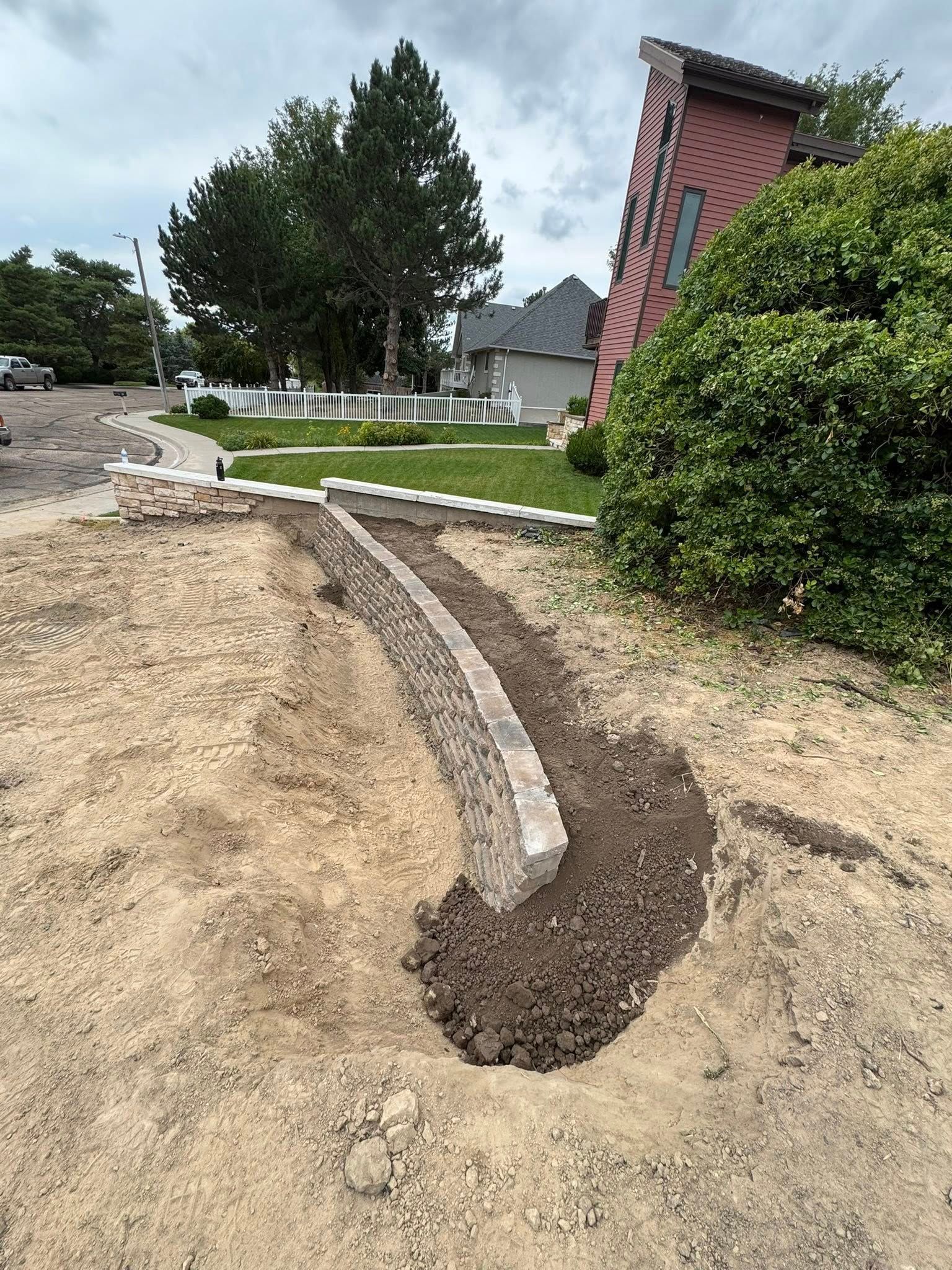 Curved concrete curb edging a dirt path beside a brick house and shrubs under construction or landscaping.