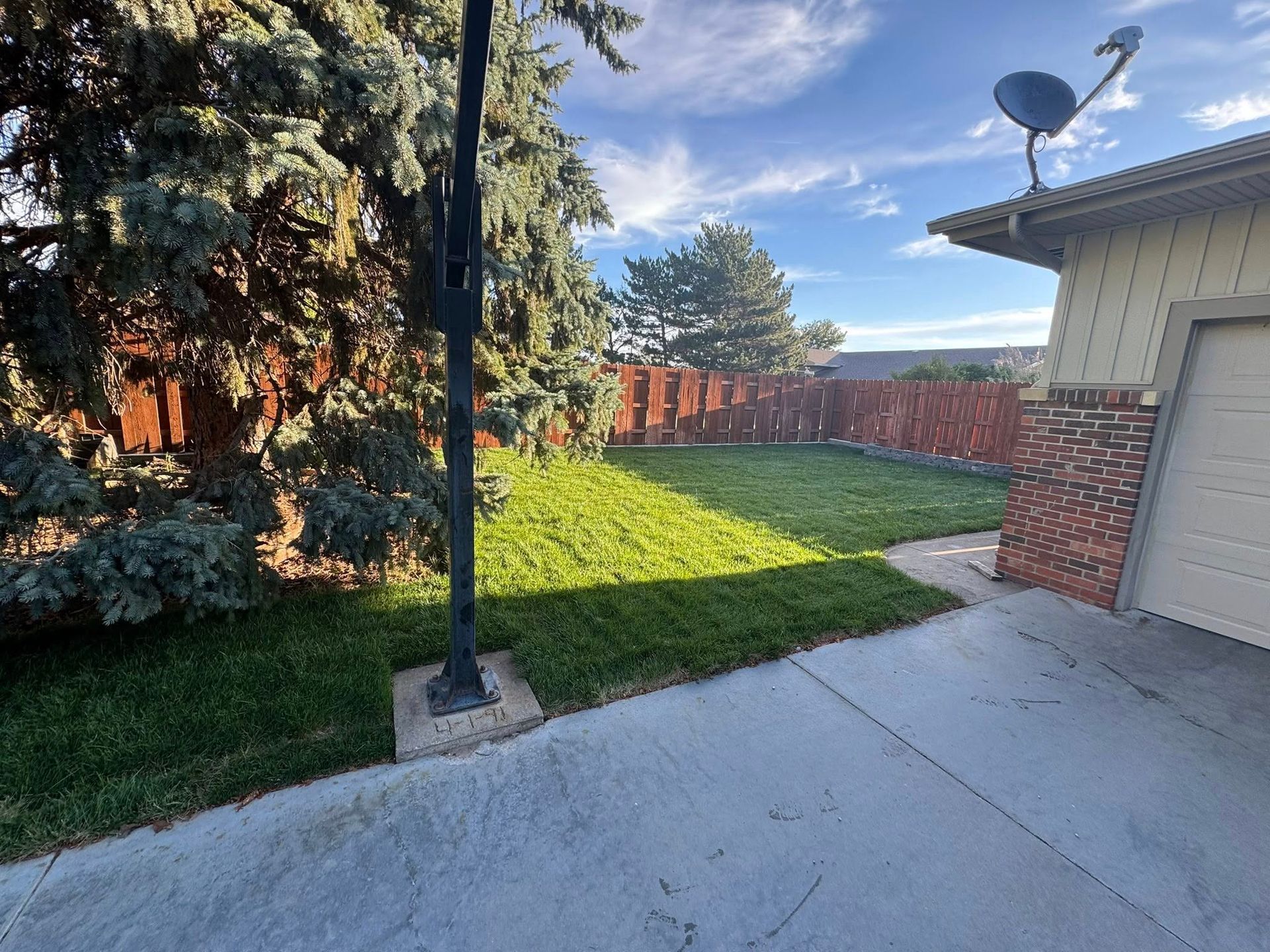 Backyard patio with concrete slab, lamp post, green lawn, trees, fence, and a house with a satellite dish