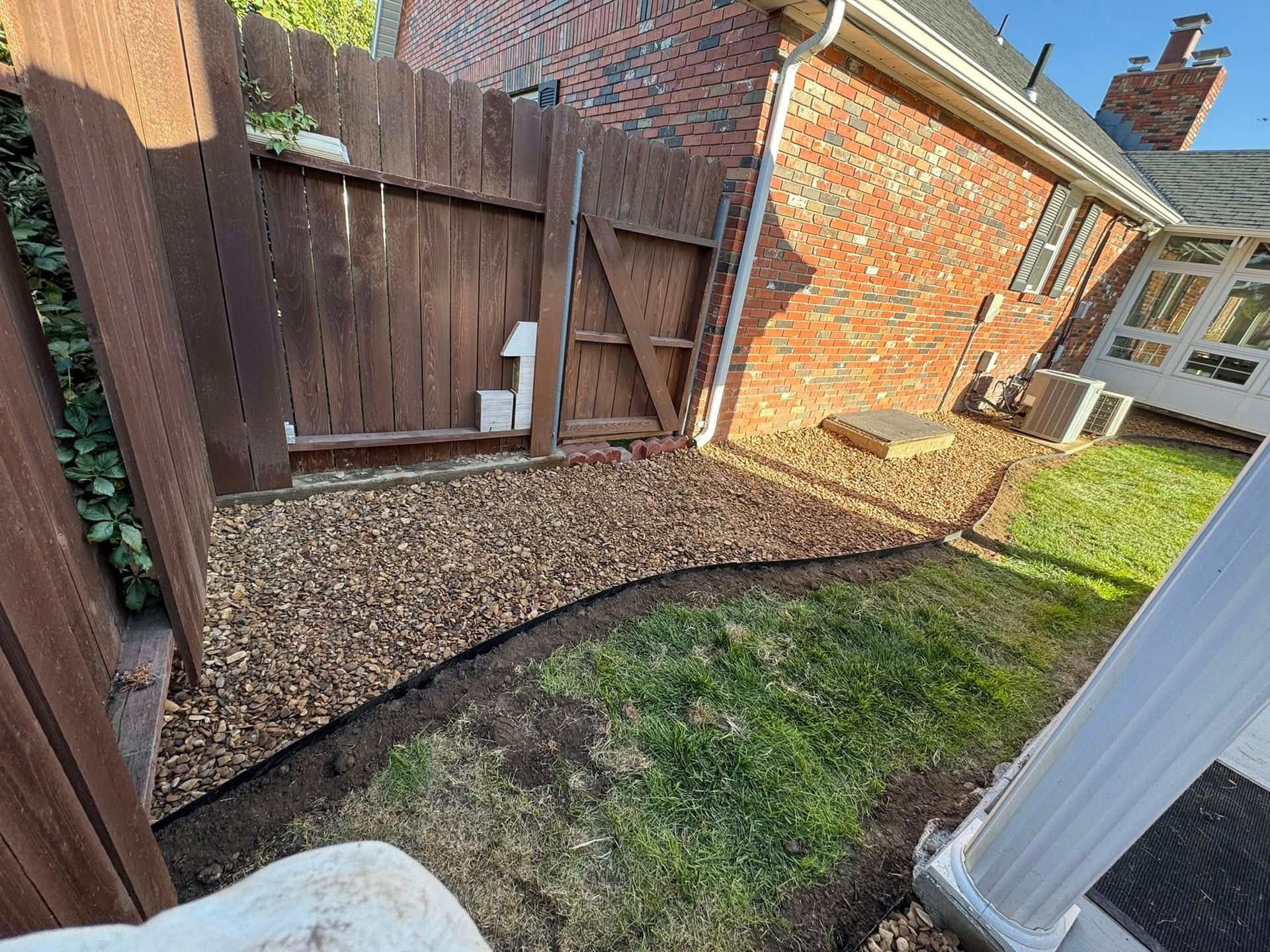 Backyard corner with wooden fence, gravel border, grass, and a brick house wall