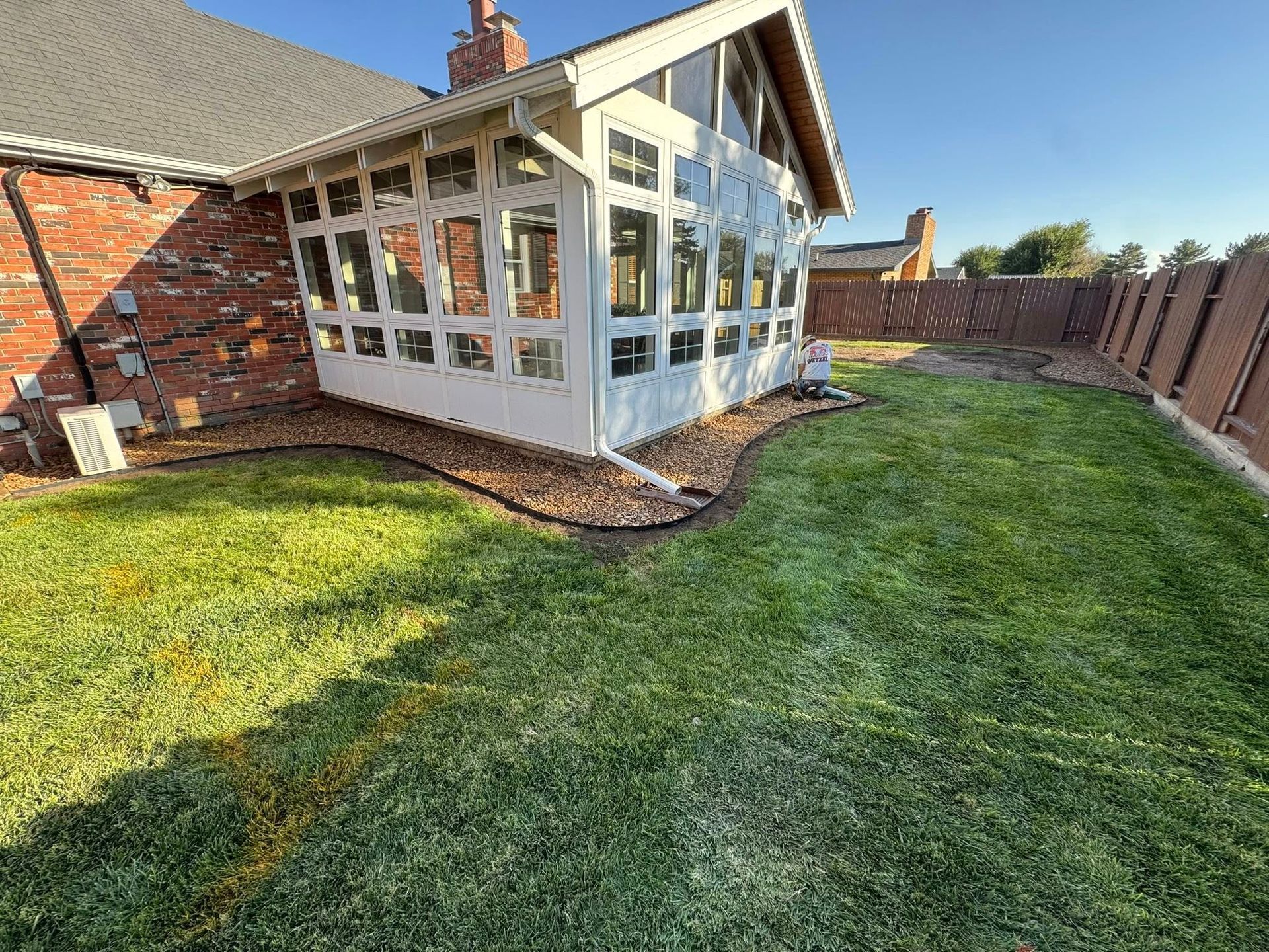 Backyard lawn beside a brick house and white sunroom with a fenced perimeter under clear sunlight