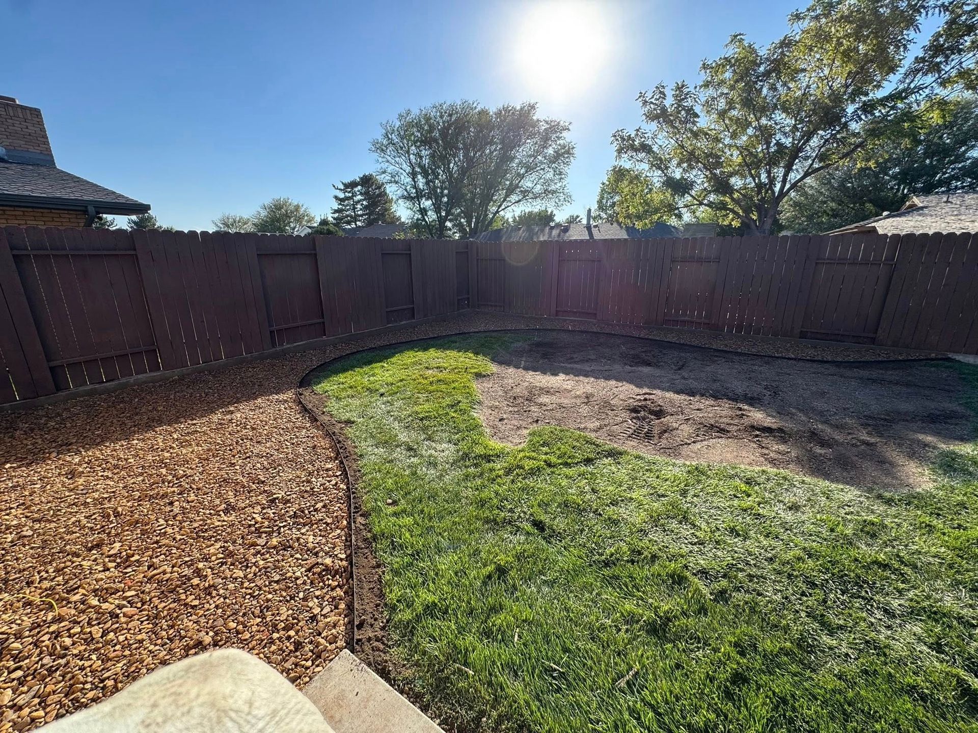 Backyard with curved mulch border, patchy grass, and a wooden fence under bright sunlight