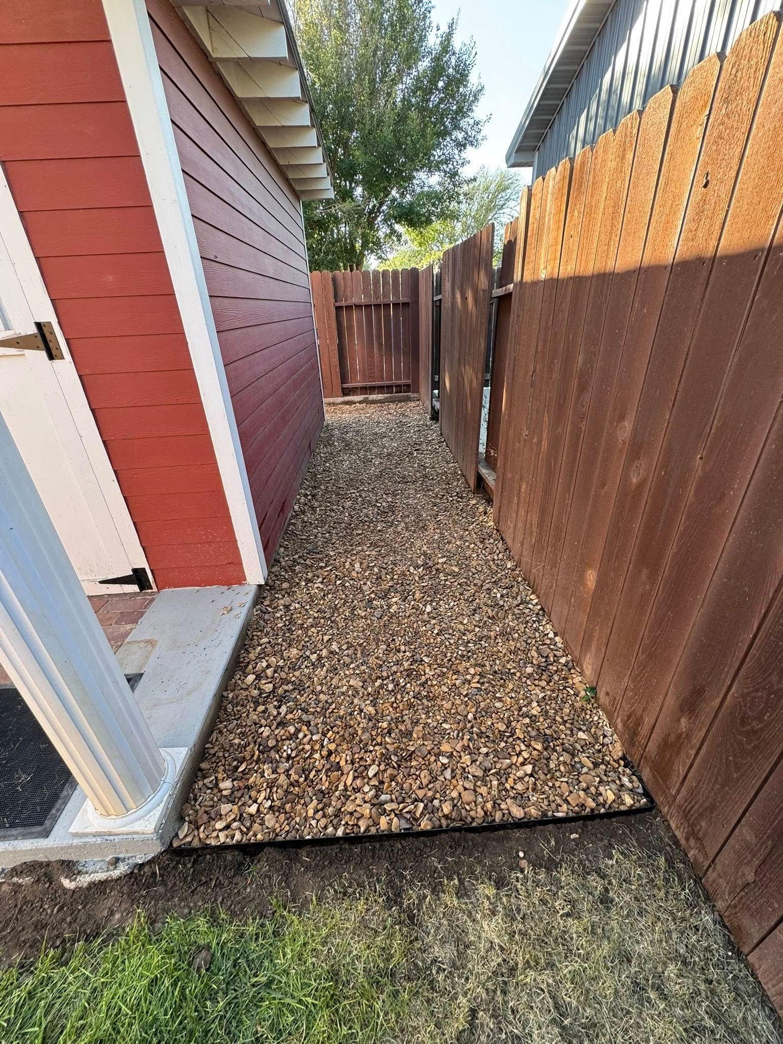 Narrow gravel path between a red house and brown fence leading to a wooden gate