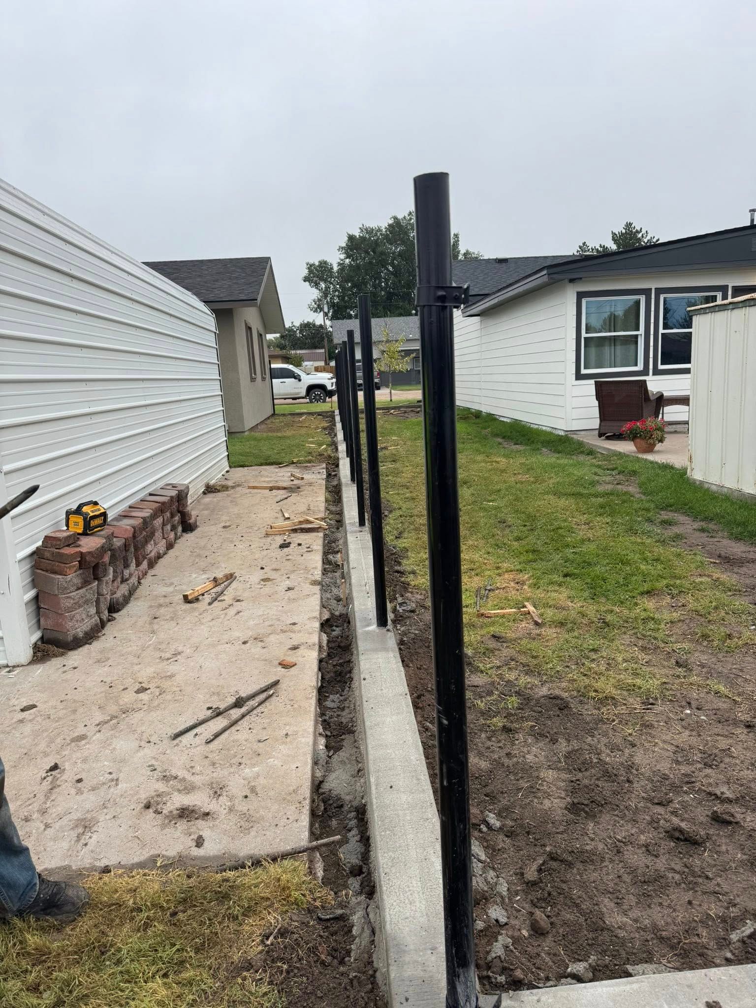 Narrow concrete walkway beside a white house, bordered by a black metal fence and gravel yard.