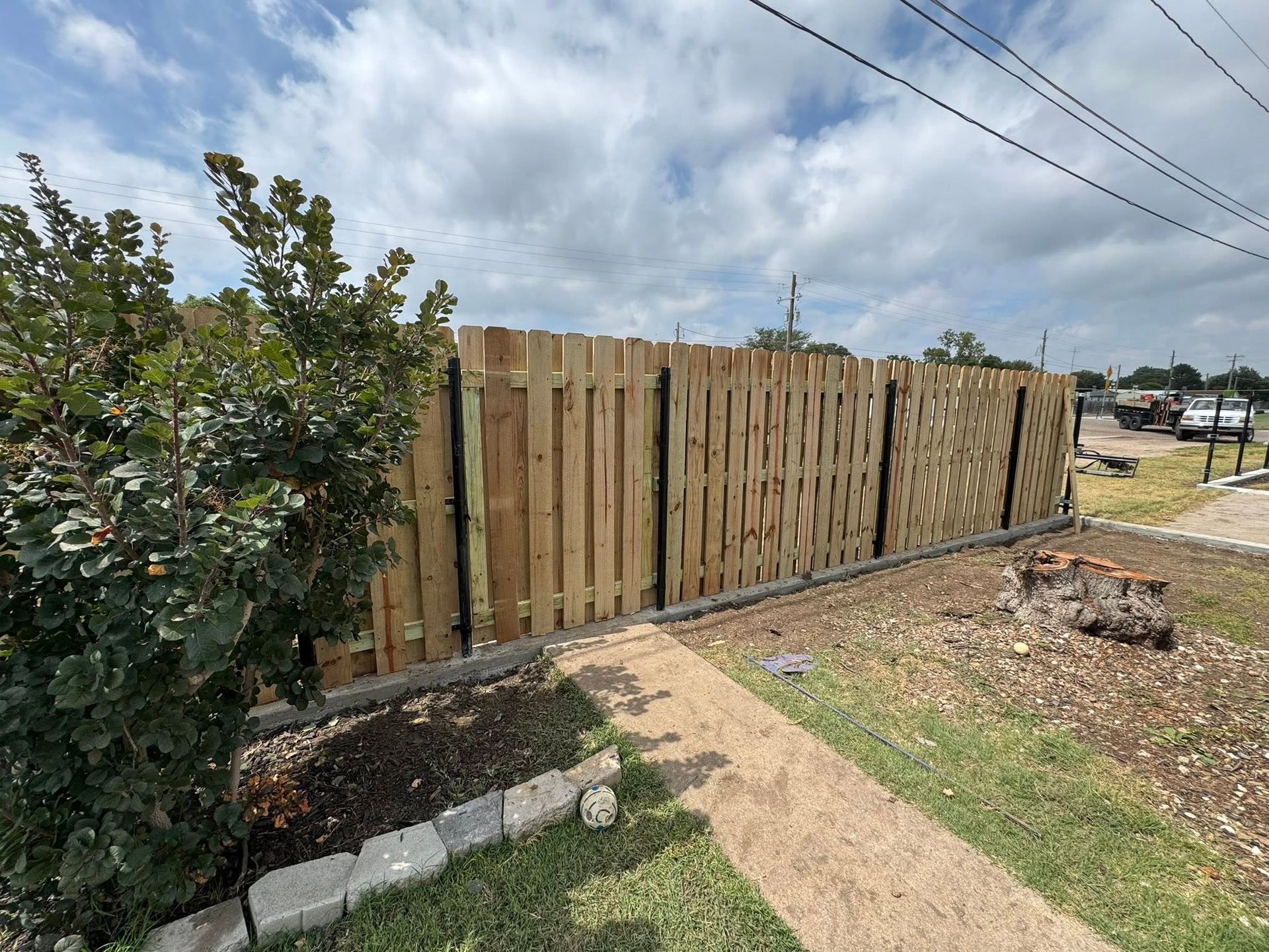 Wooden fence along a dirt path beside shrubs under a cloudy sky
