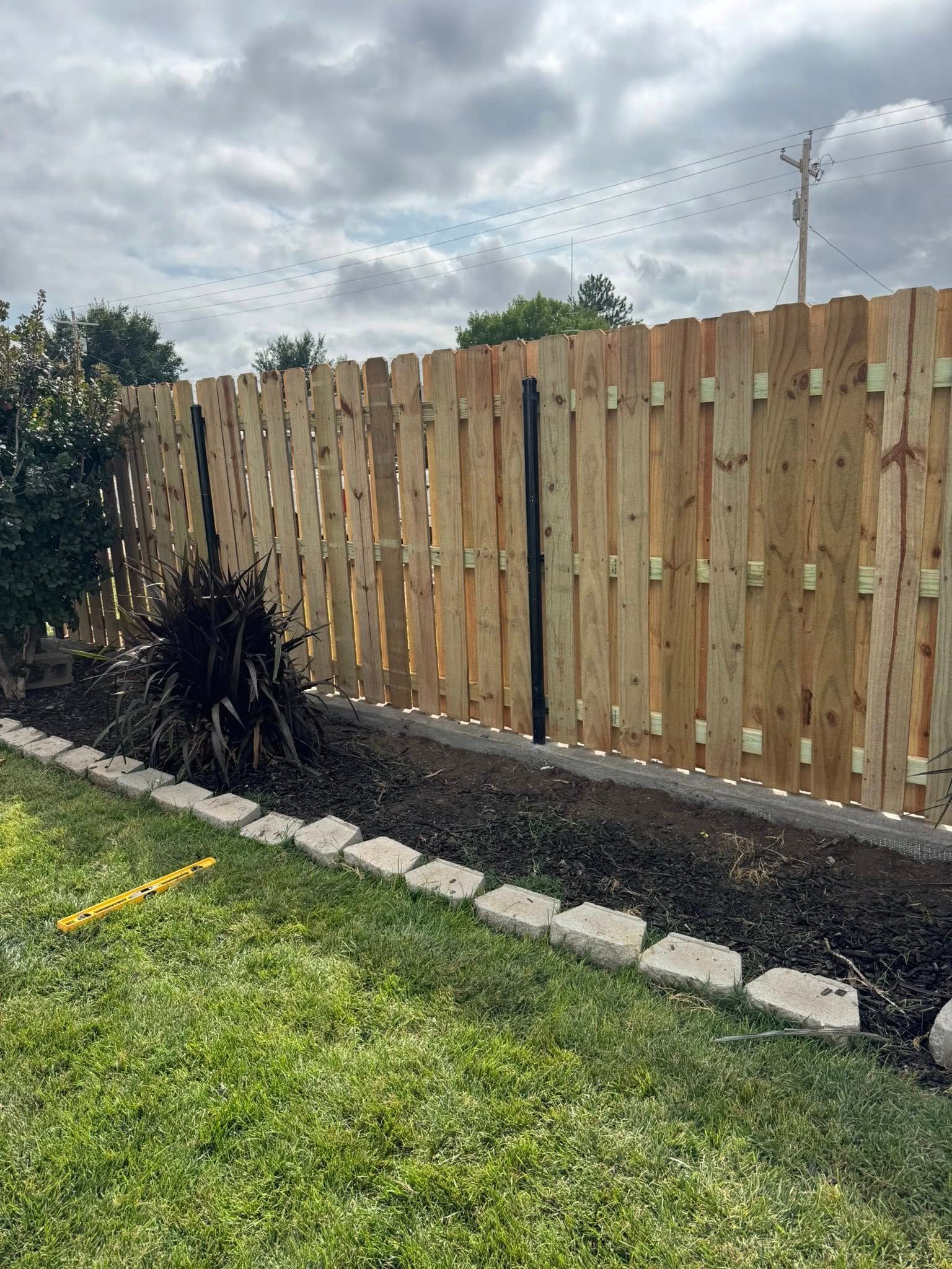 Backyard fence with dark mulch bed, small shrub, and green lawn under a cloudy sky