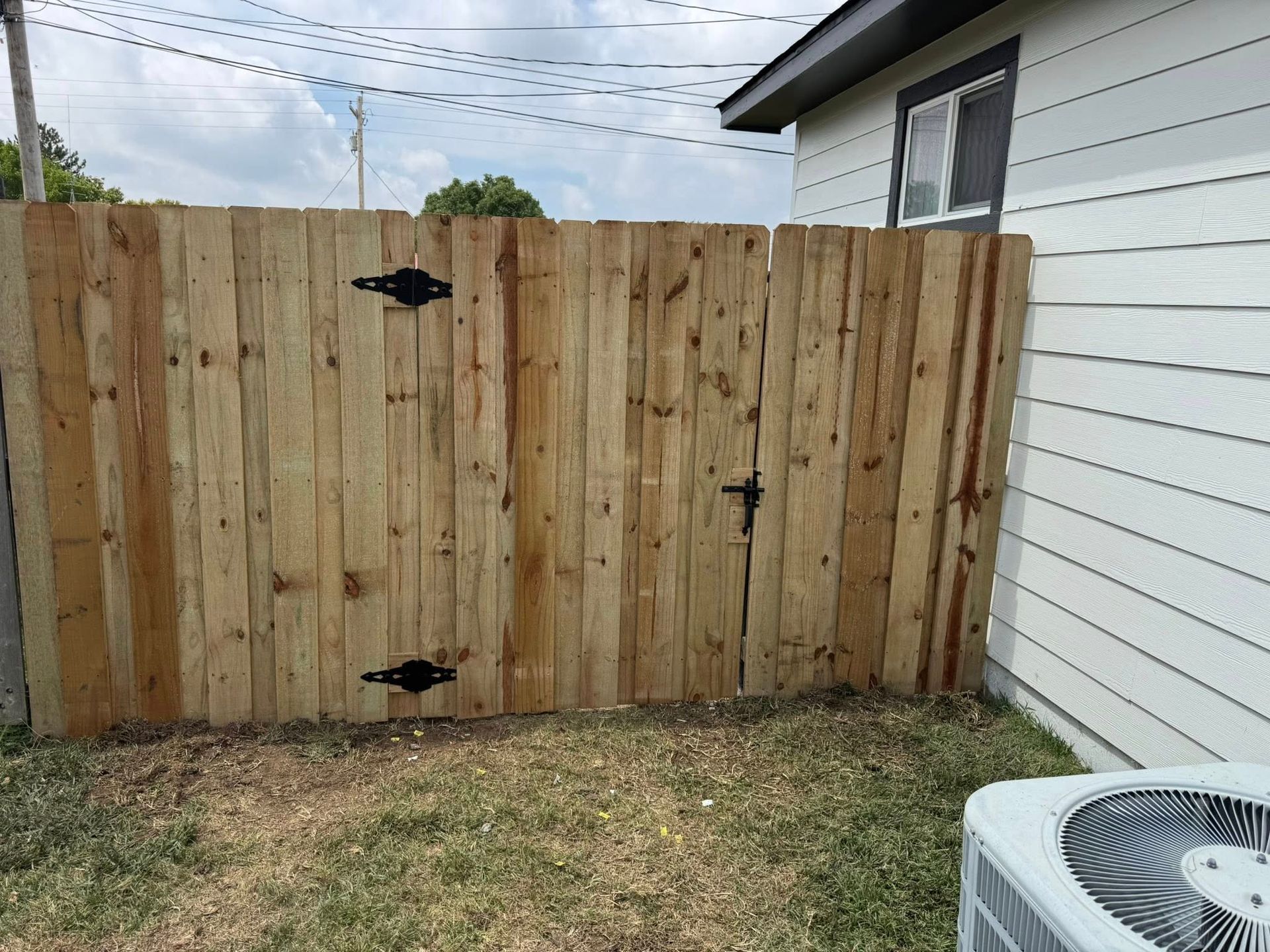Wooden backyard gate beside white house siding, with grass and an air conditioner visible.