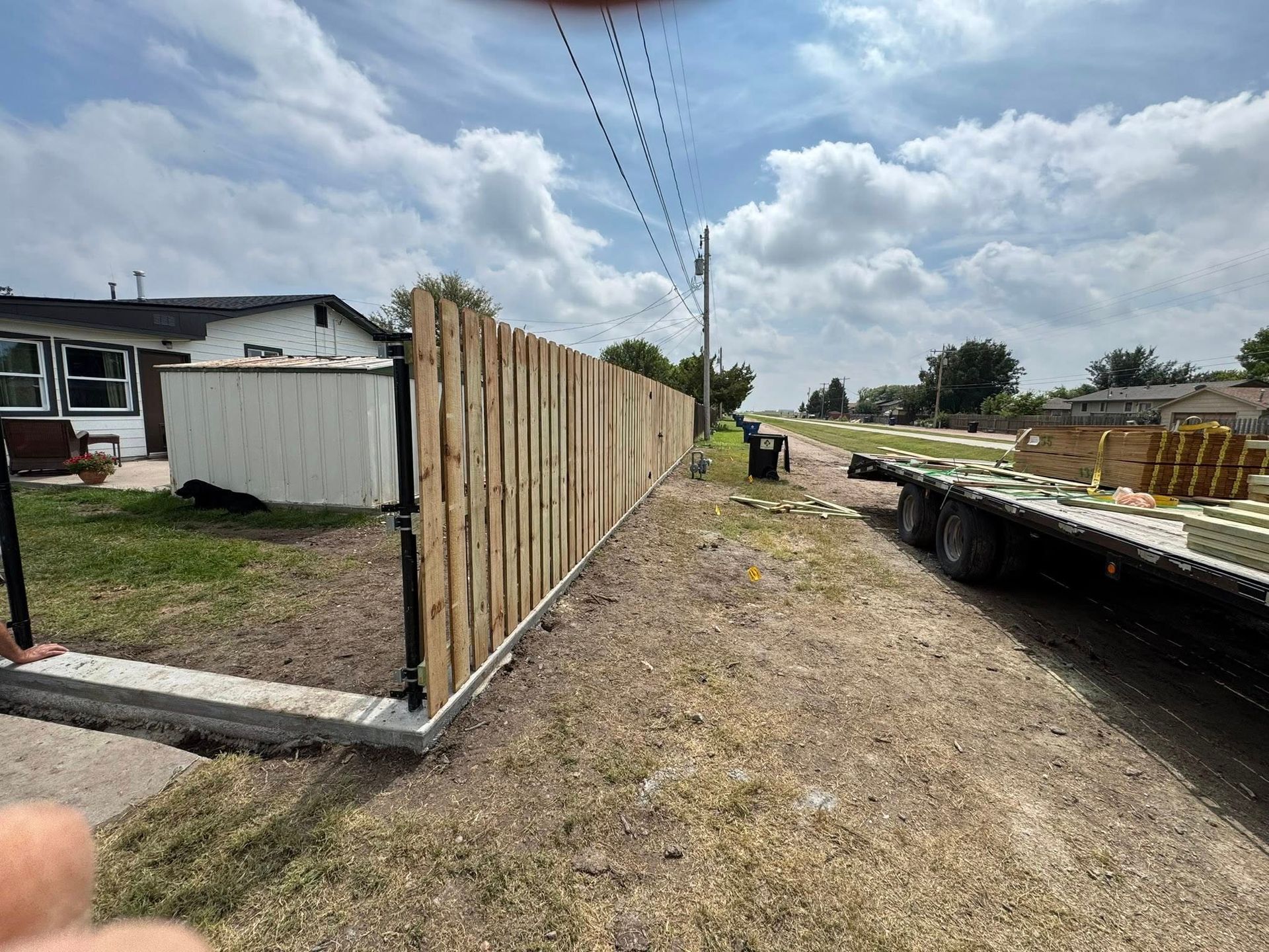 Construction site with wooden fence, gravel path, and flatbed trailer under cloudy sky